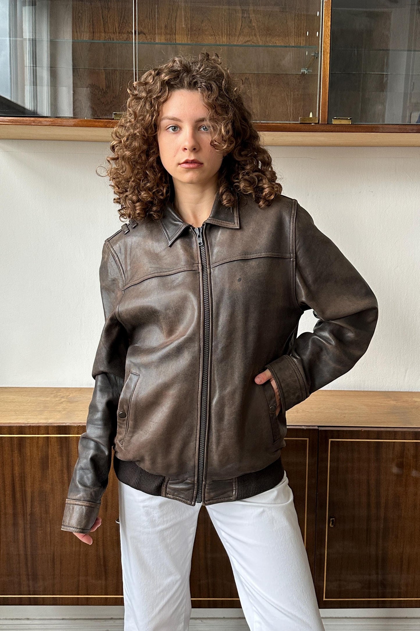 A young woman with curly hair wearing a brown leather jacket stands in front of a wooden cabinet.