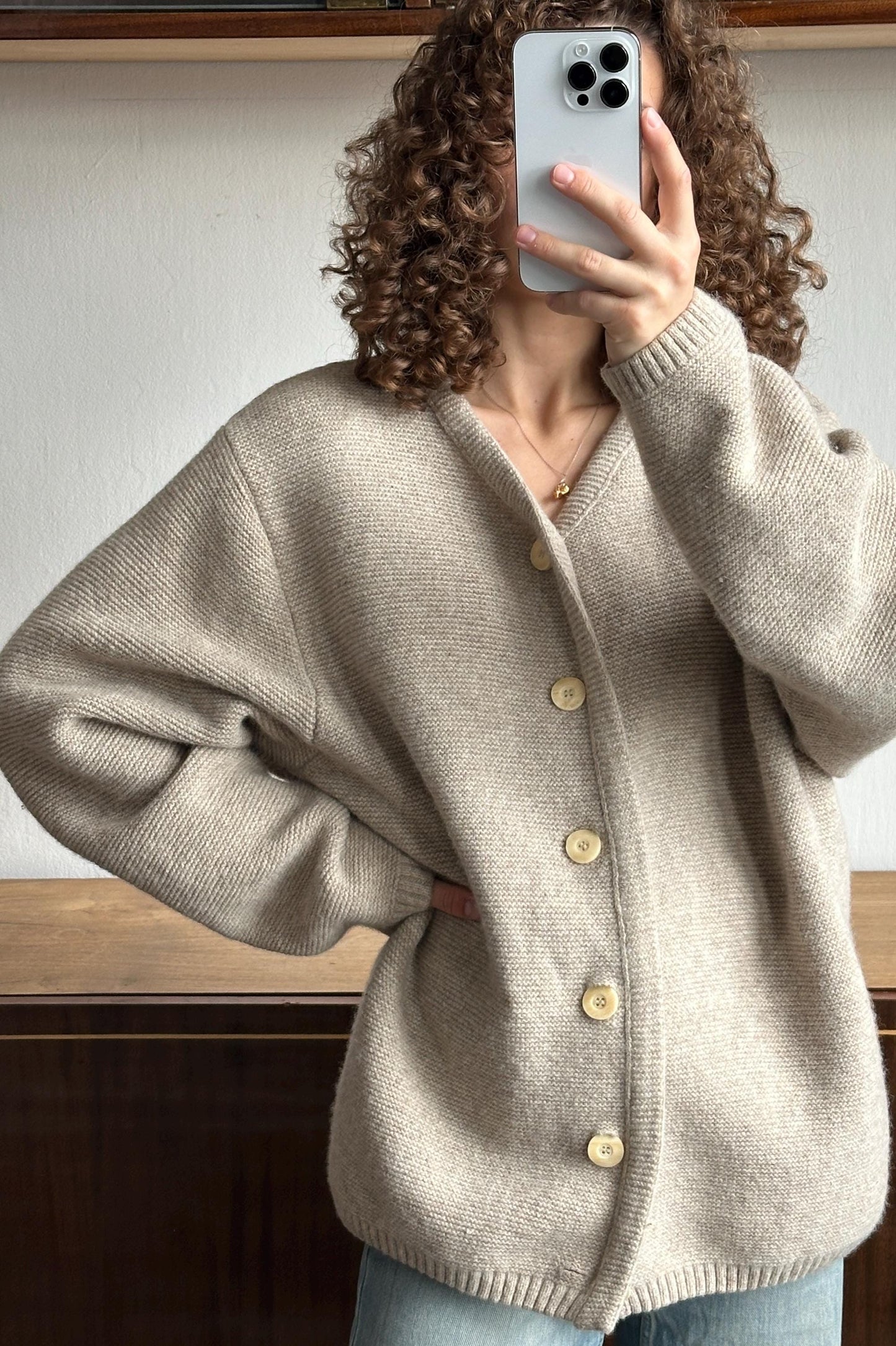A person with curly hair is taking a selfie with a smartphone, wearing a beige cardigan.