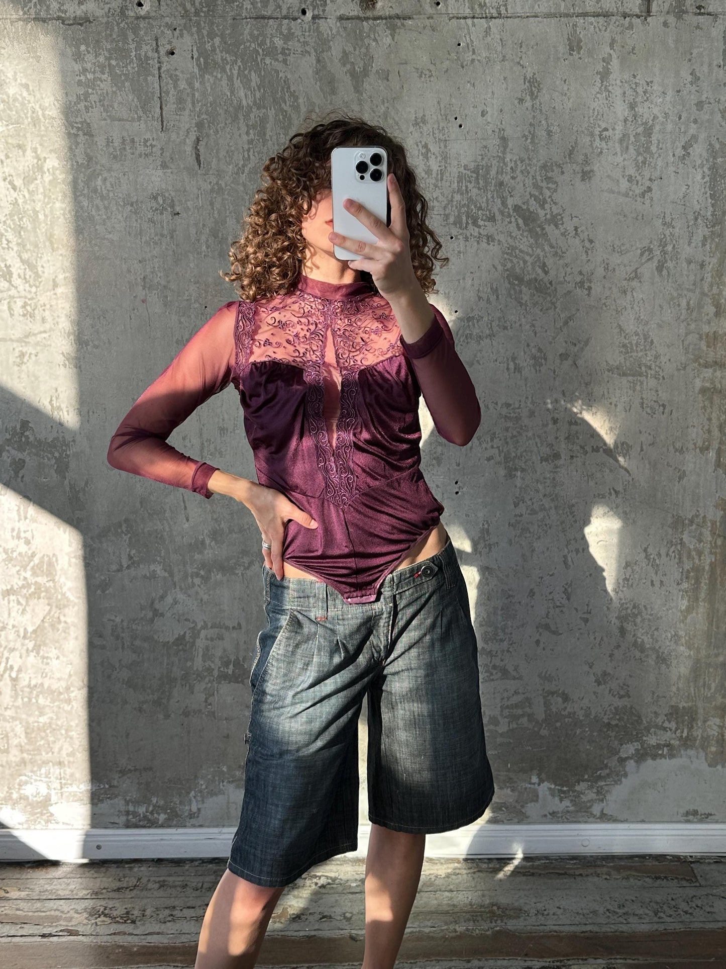A woman is taking a selfie with a white phone, wearing a purple top and blue jeans, against a concrete wall.