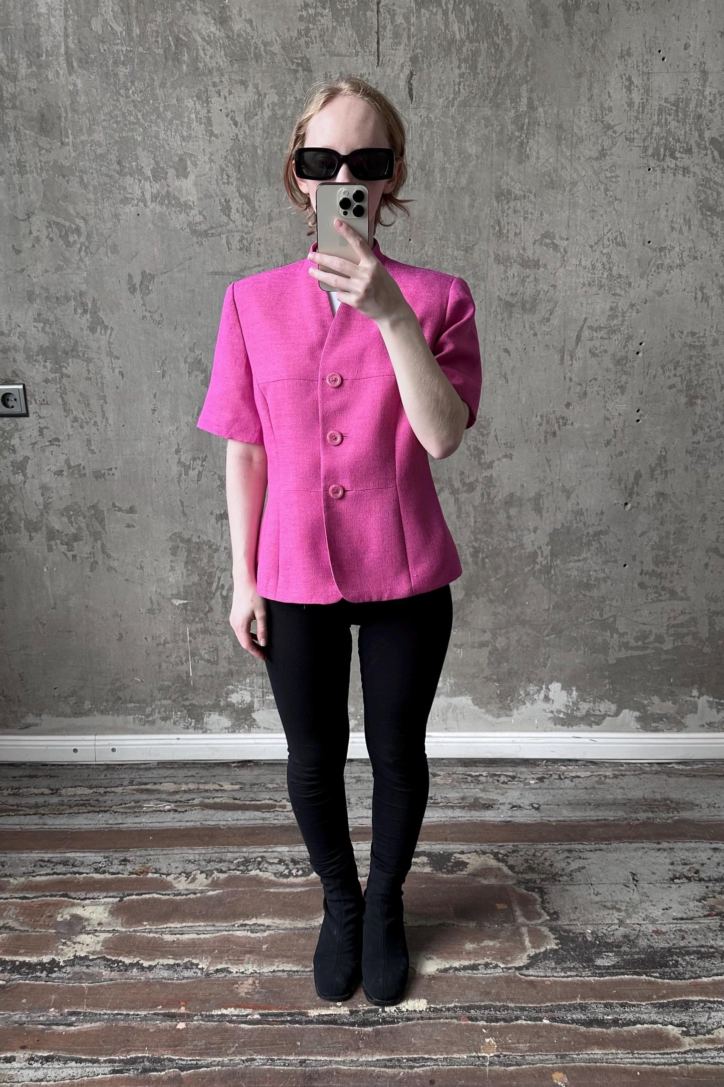A woman wearing a pink jacket and sunglasses is taking a selfie with a smartphone in front of a concrete wall.