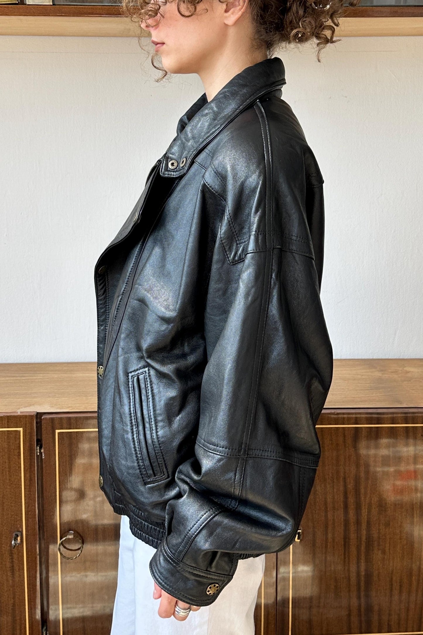 A person wearing a black leather jacket stands in front of a wooden cabinet.