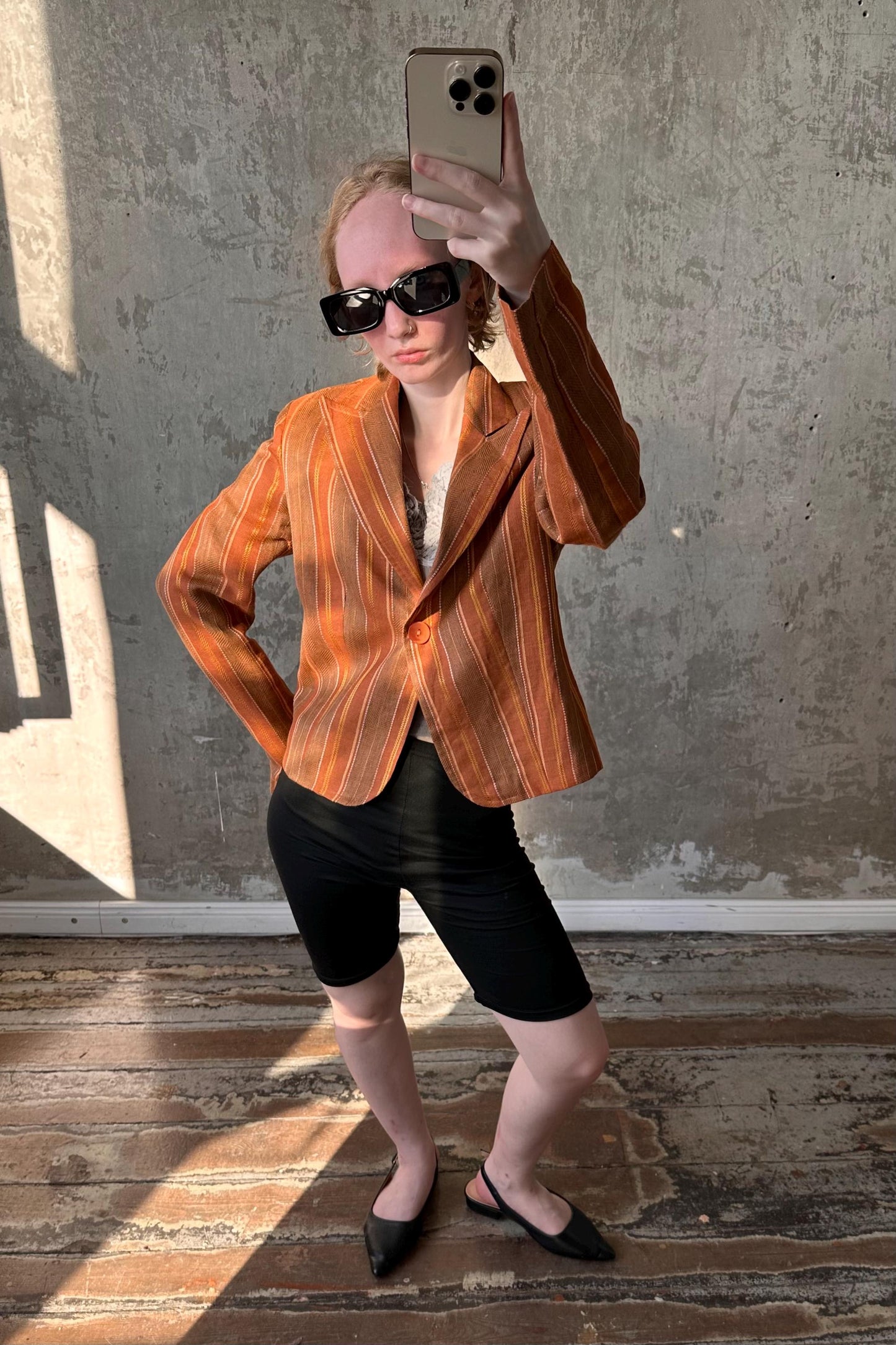 A person wearing a striped orange jacket, sunglasses, and black shorts is taking a selfie with a smartphone.
