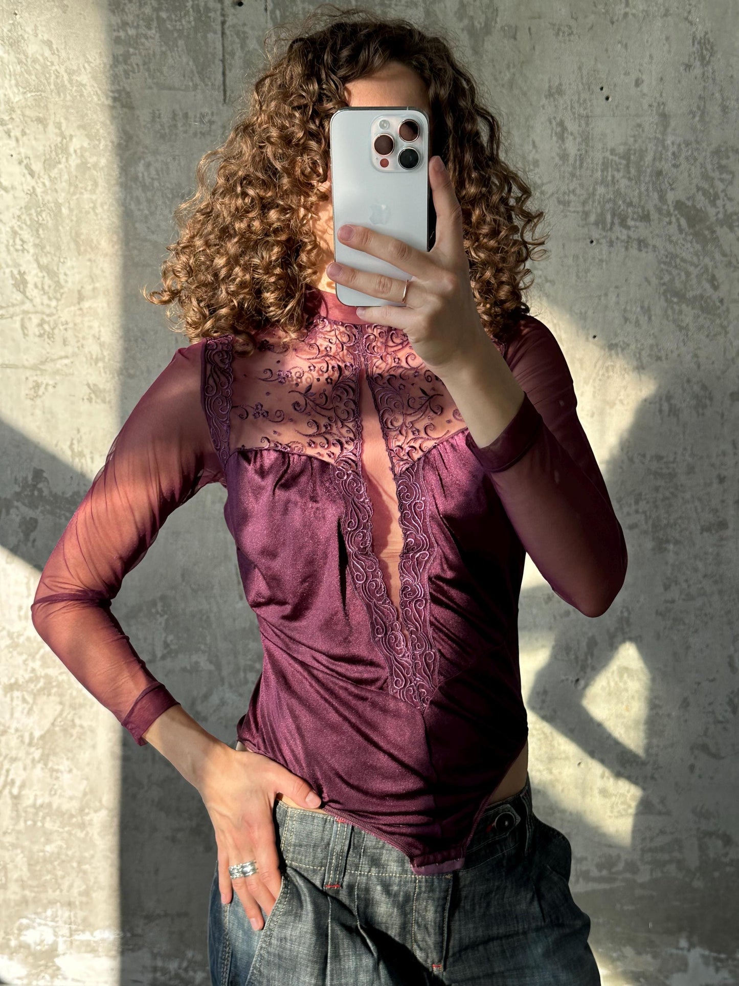 A woman with curly hair is taking a selfie with her iPhone, wearing a purple top with lace detailing and jeans.