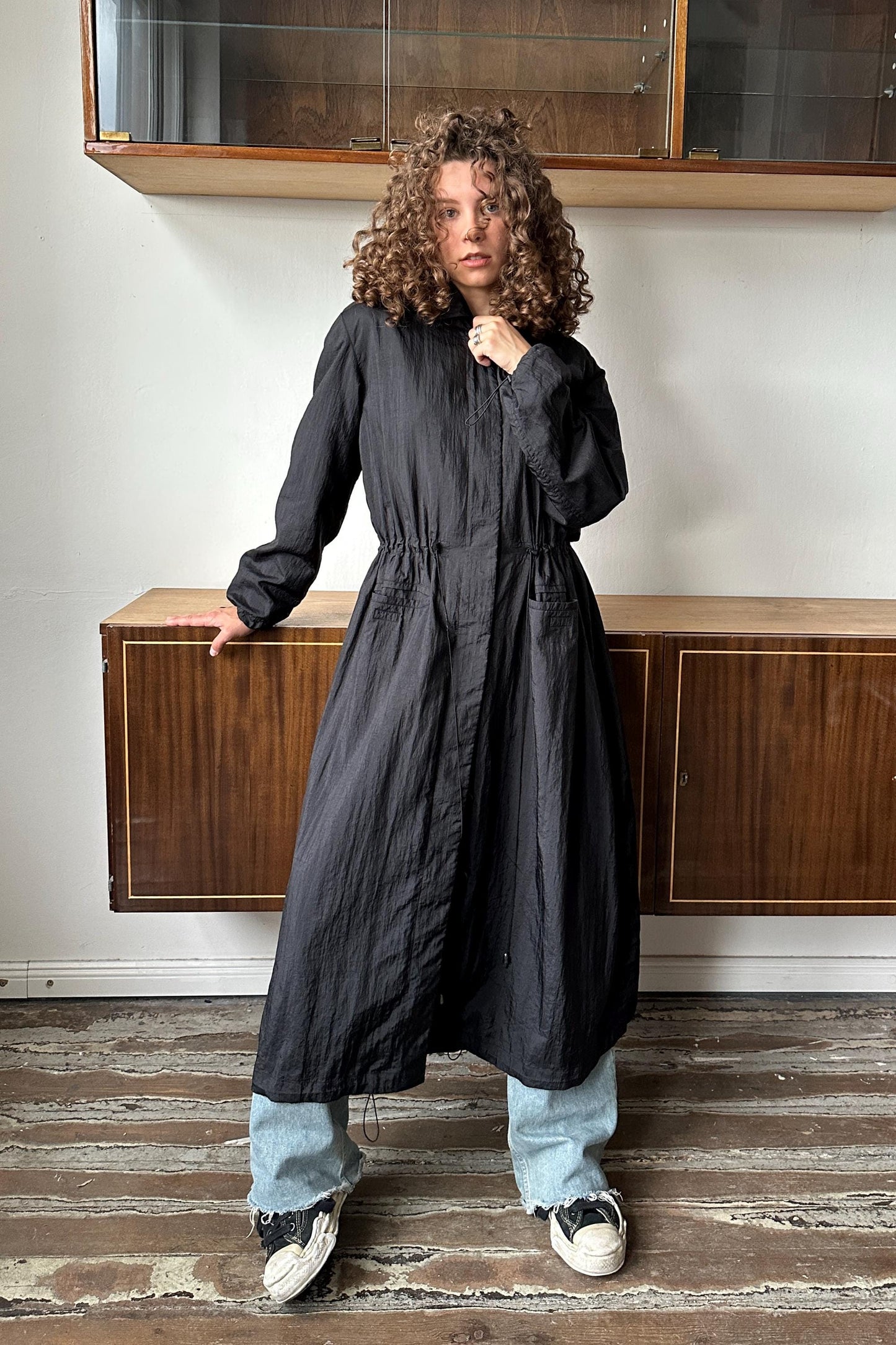 A woman with curly hair wearing a long black coat stands in a room with wooden cabinets and a glass door.