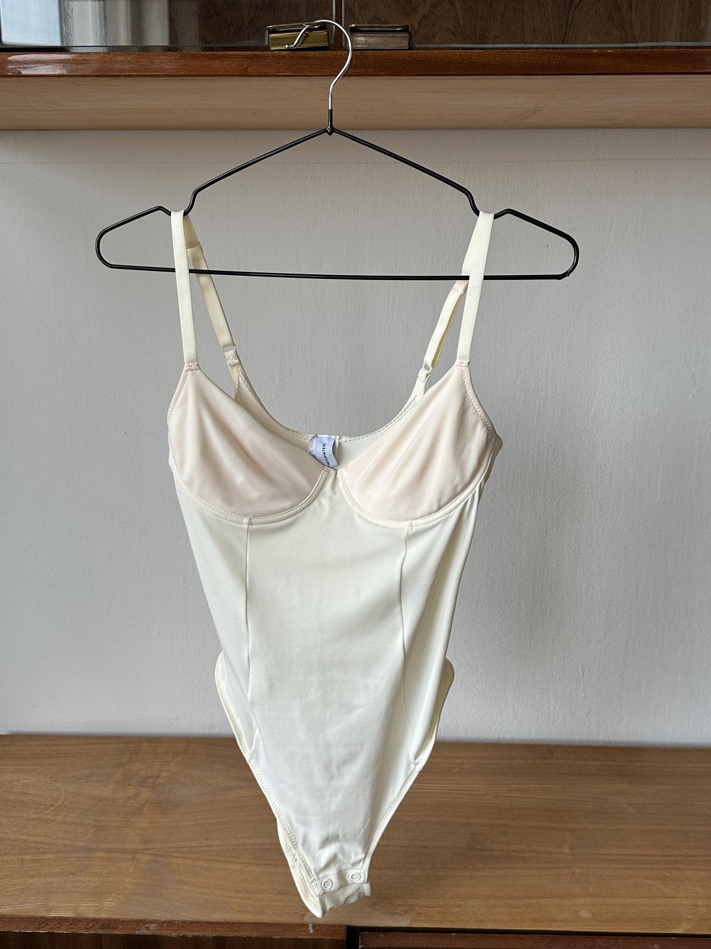 A white one-piece swimsuit is hanging on a black hanger on a wooden shelf.