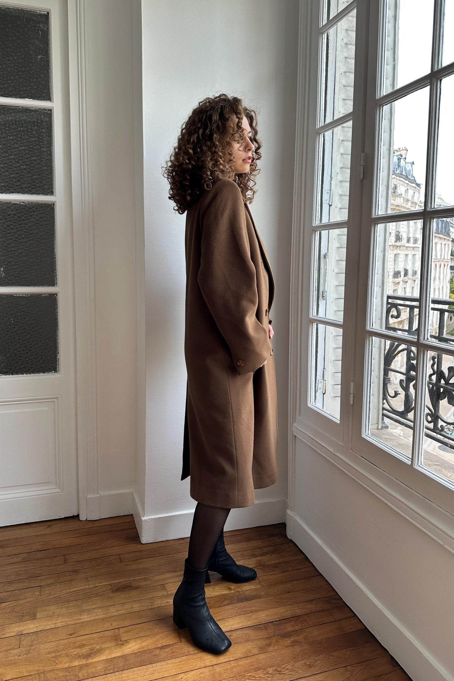 A woman with curly hair stands in a room, wearing a brown coat and black boots, looking out a window.