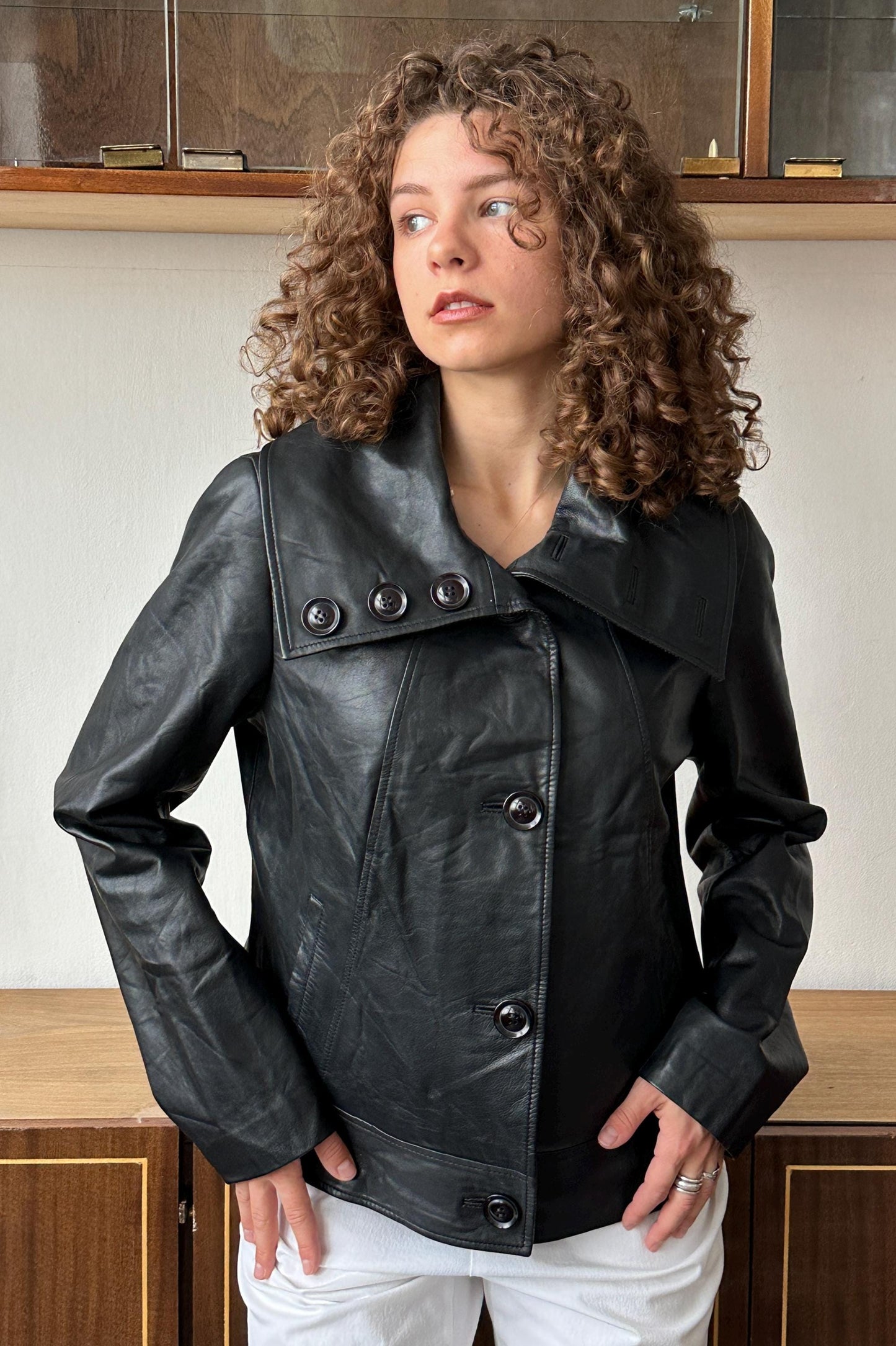 A woman with curly hair wearing a black leather jacket stands in front of a wooden cabinet.