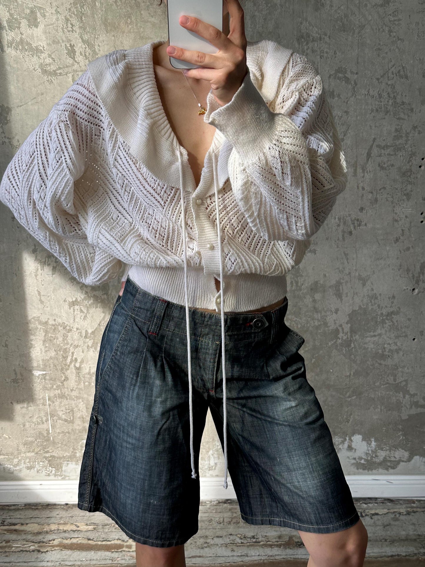 A person is taking a selfie with a white cardigan and blue denim shorts, holding a white object in their hand.
