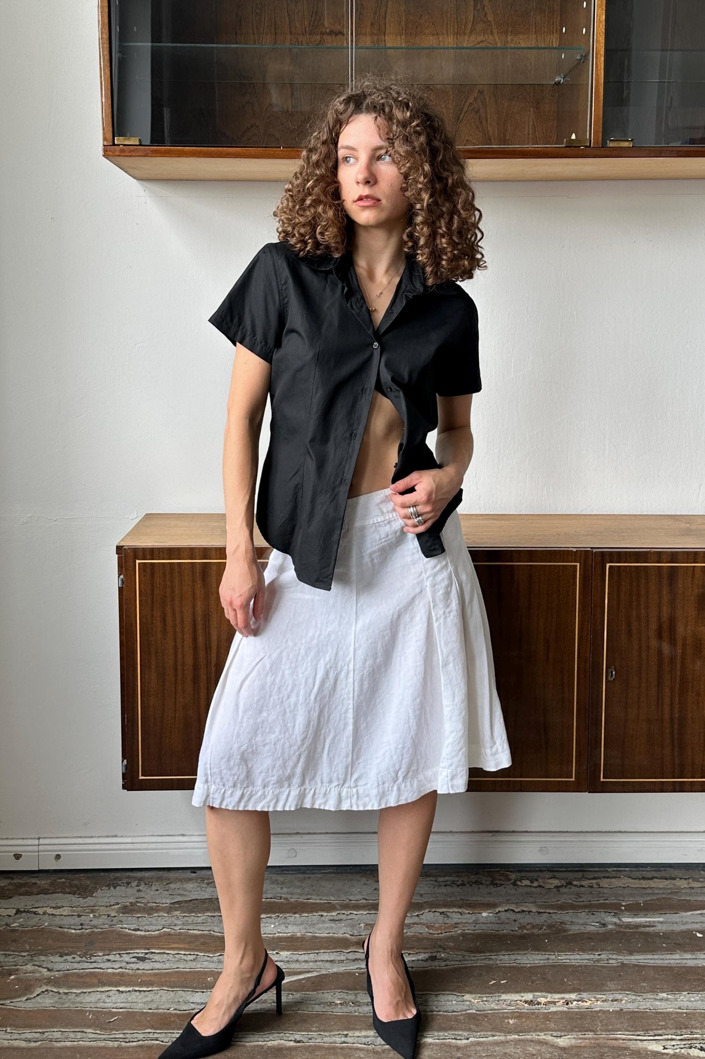 A woman with curly hair wearing a black shirt and white skirt stands in front of a wooden cabinet.