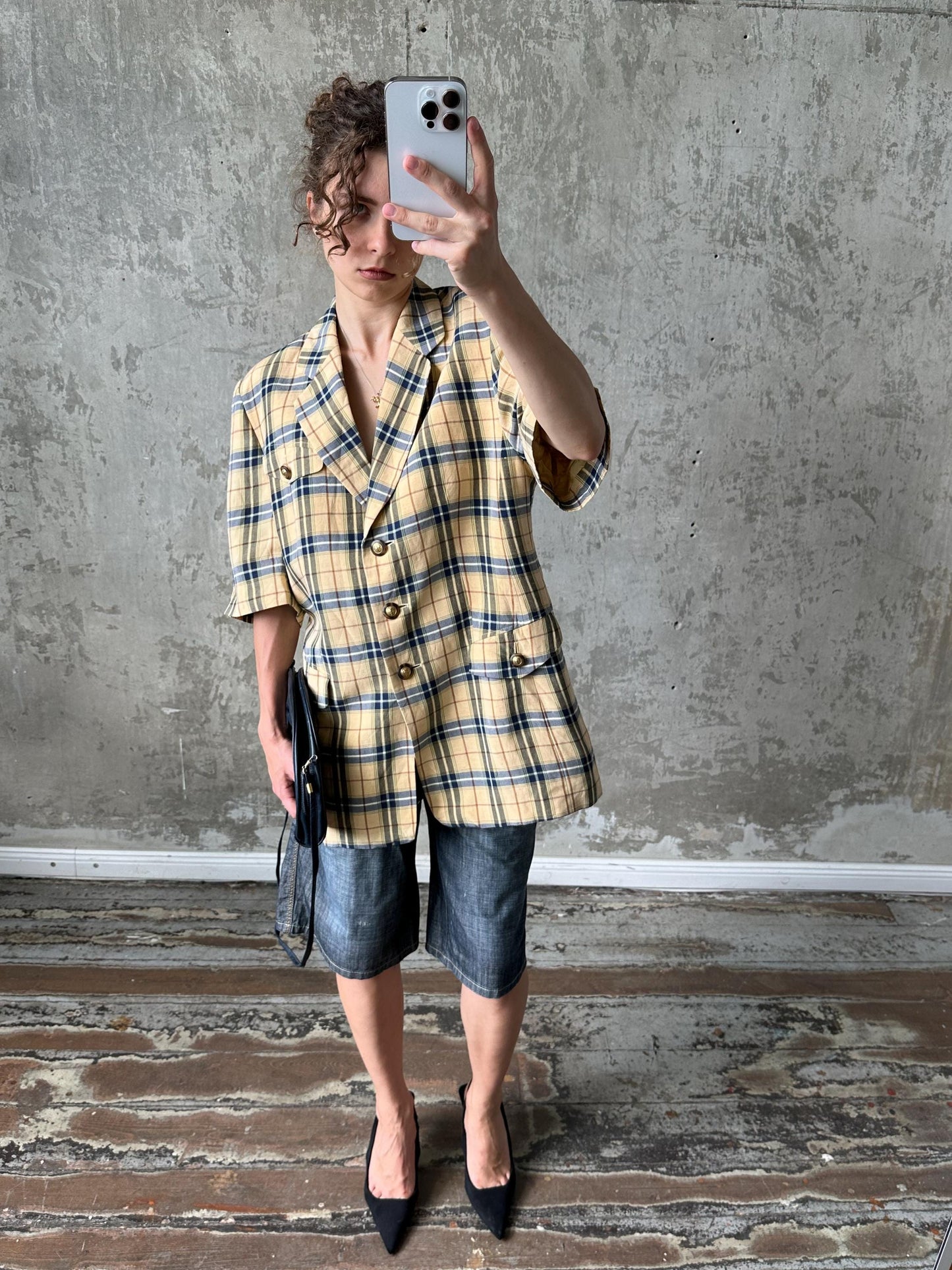 A person is taking a selfie with a smartphone, wearing a yellow and black plaid jacket, denim shorts, and black heels.