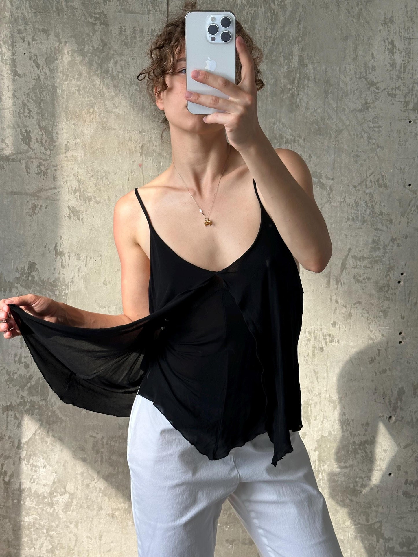 A woman is taking a selfie with her iPhone, holding the device up to her face and covering her mouth. She is wearing a black top and white pants.