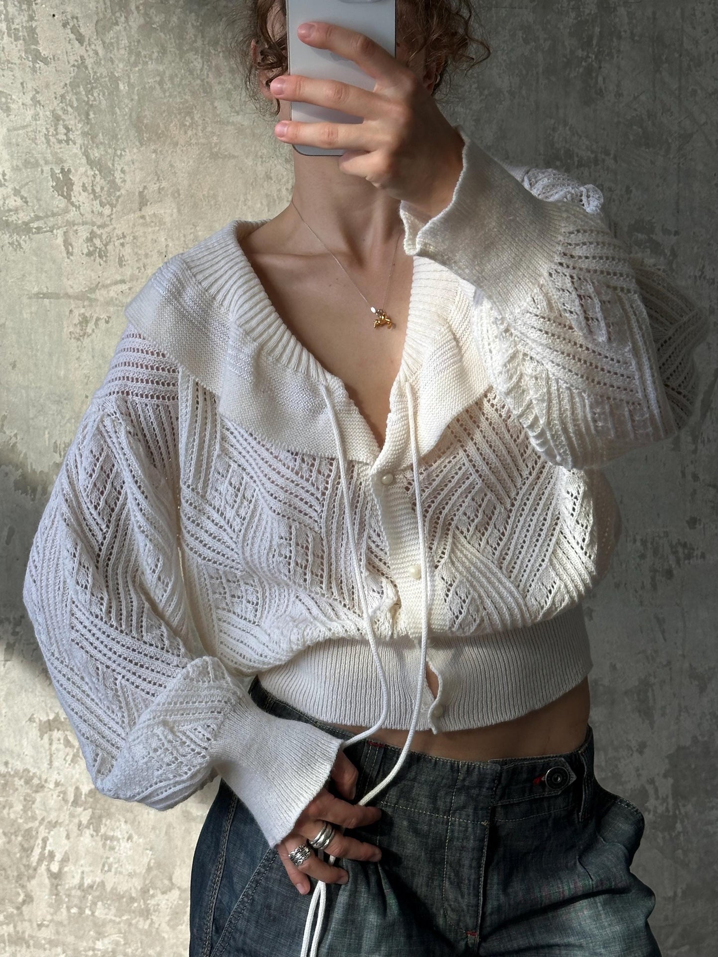 A person is taking a selfie with a white cardigan and jeans, holding a phone up to their face.