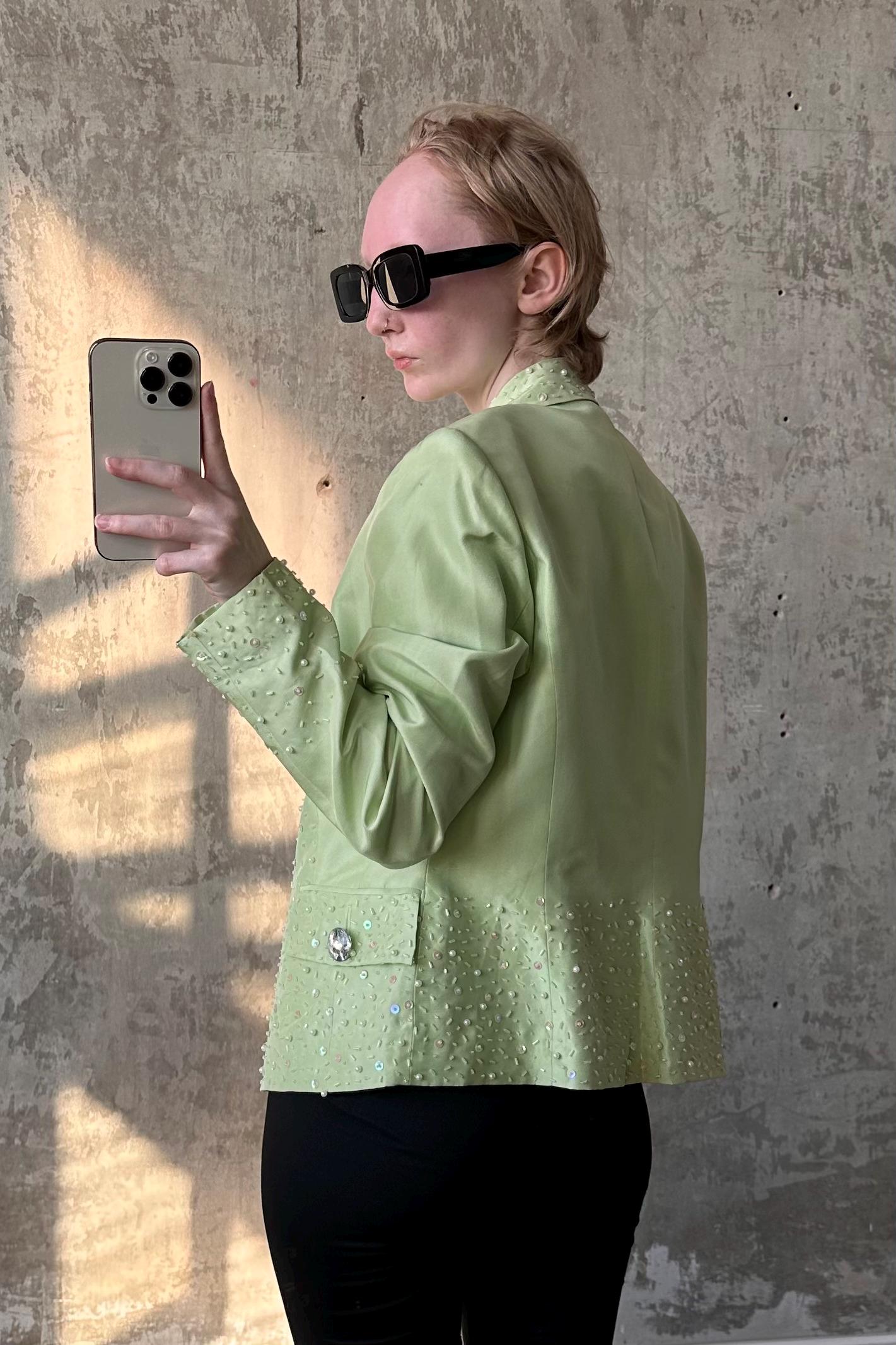 A person wearing a green jacket with embellishments and sunglasses is taking a selfie with a smartphone.