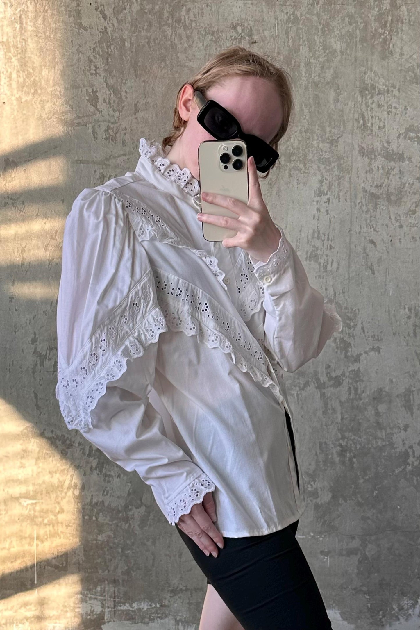 A person is taking a selfie with a smartphone, wearing a white lace blouse and sunglasses.