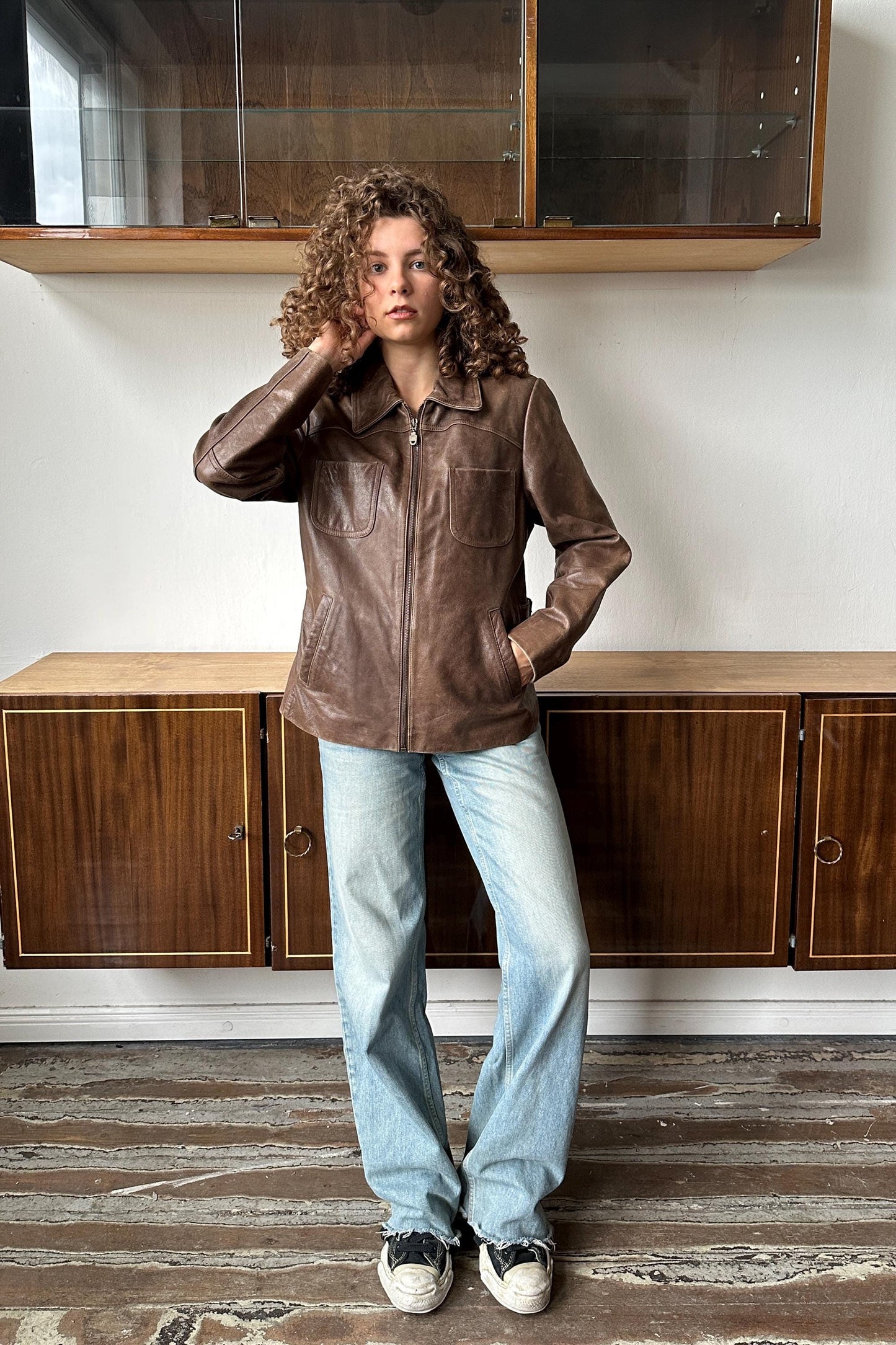 A woman with curly hair stands in a room, wearing a brown leather jacket and blue jeans.