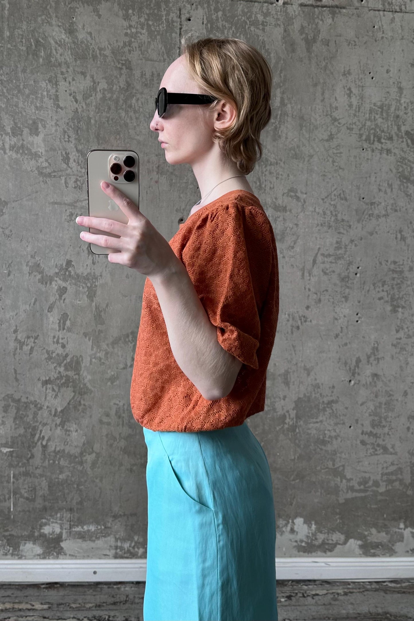 A woman in an orange top and blue skirt is taking a selfie with a smartphone, holding the device up to her face and looking at the screen.