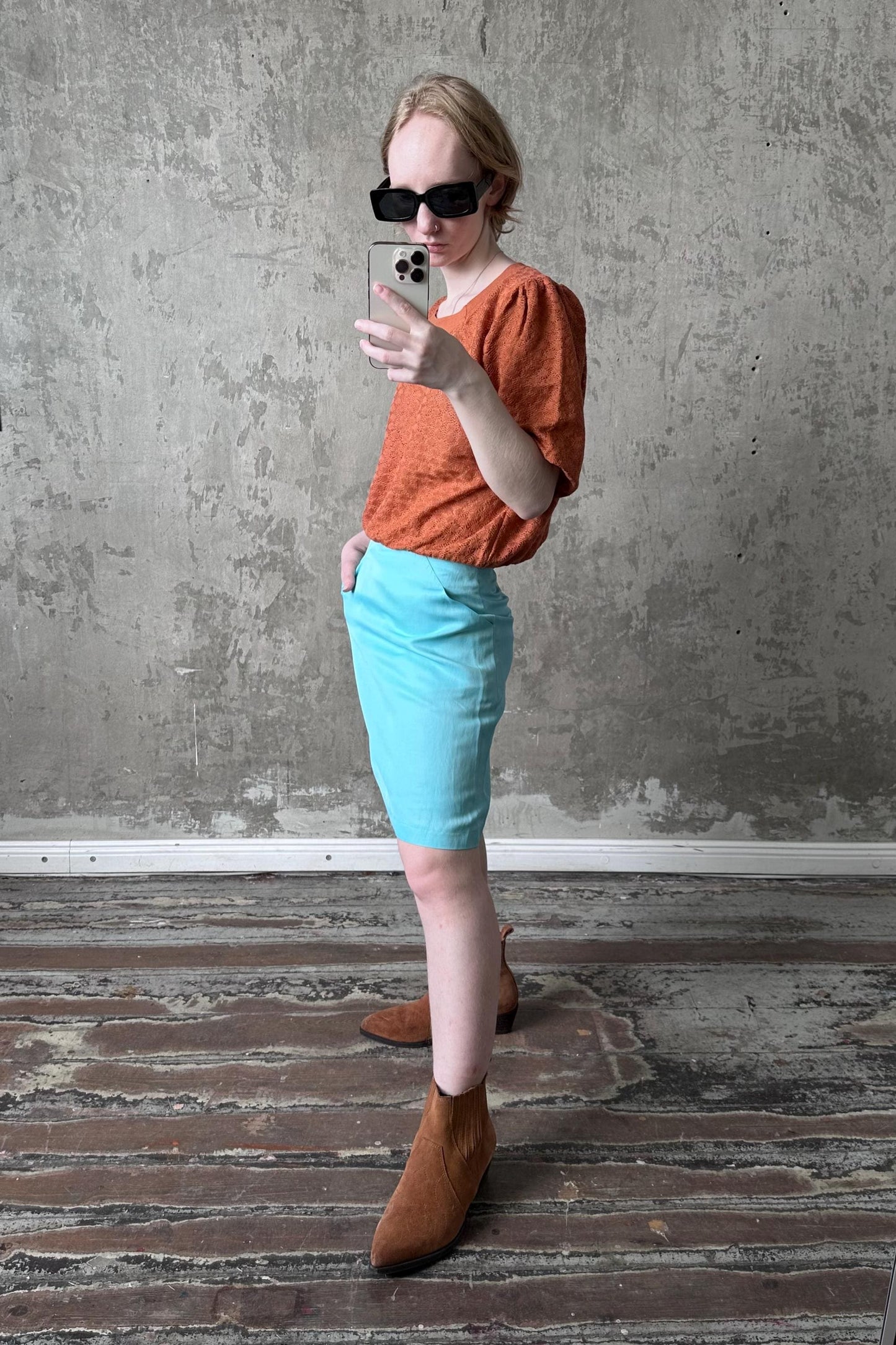 A person is taking a selfie with a smartphone, wearing a red top, blue skirt, and brown boots, standing in front of a concrete wall.