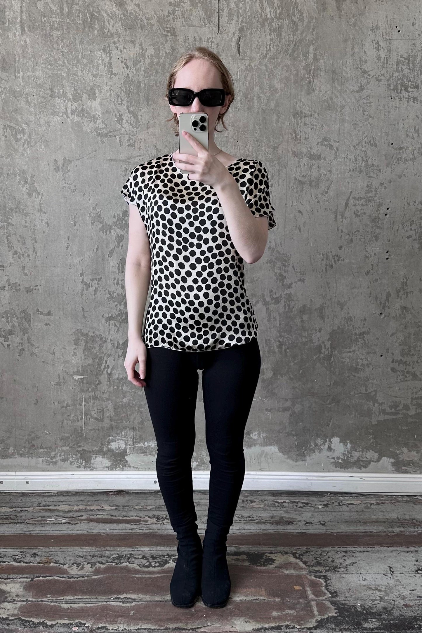 A woman wearing a black and white polka dot blouse and black leggings is taking a selfie with a smartphone against a concrete wall.