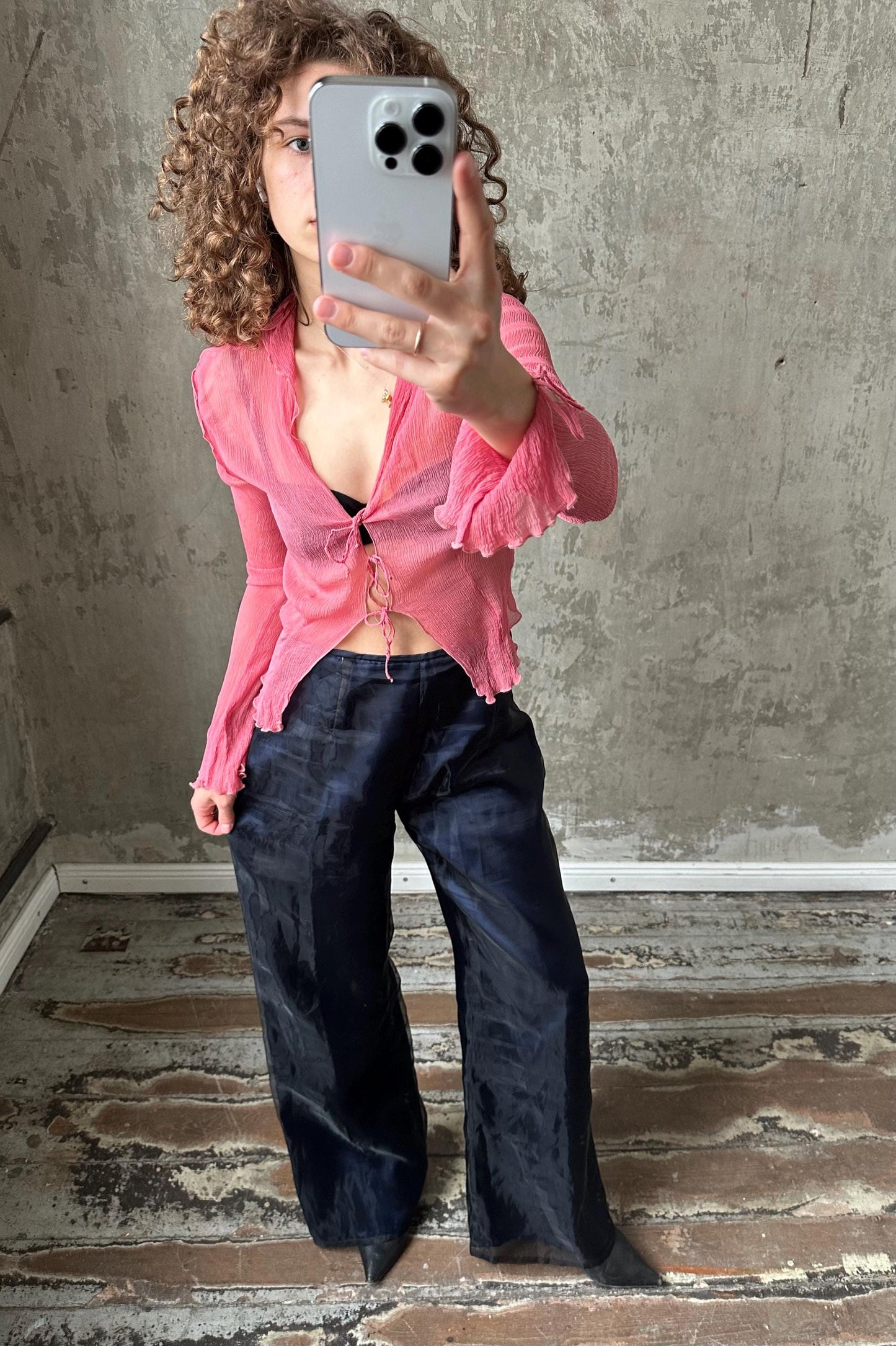 A woman with curly hair is taking a selfie with a smartphone, wearing a pink blouse and black pants.