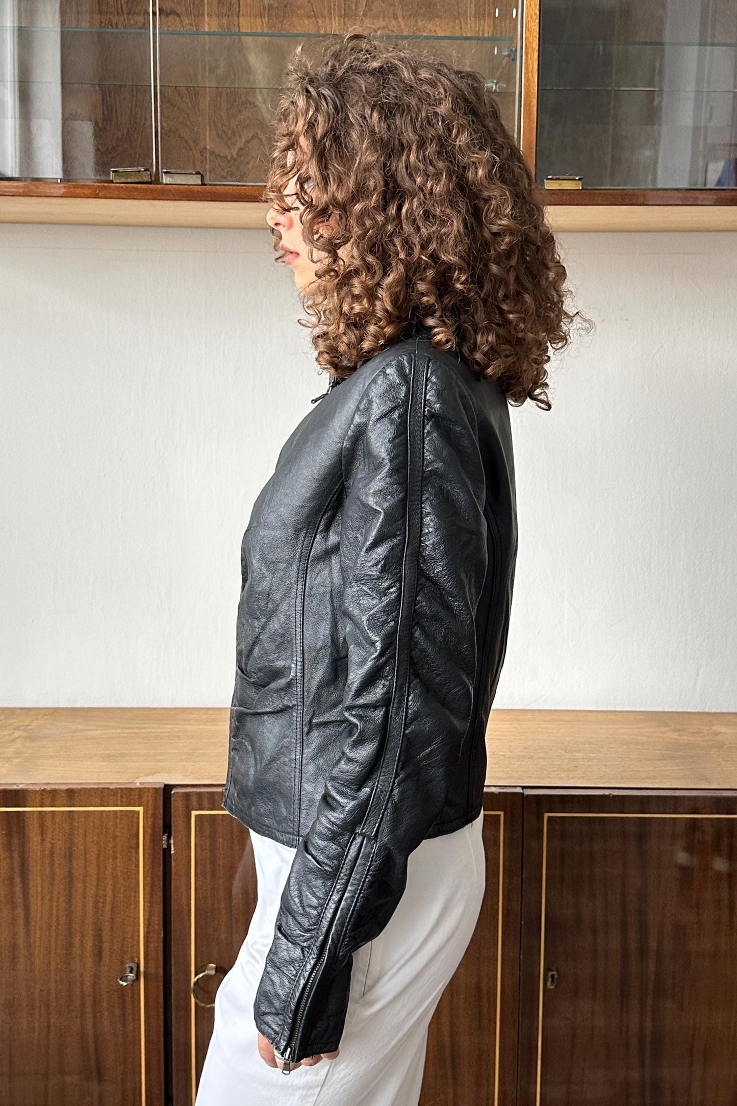 A woman with curly hair wearing a black leather jacket stands in front of a wooden cabinet.