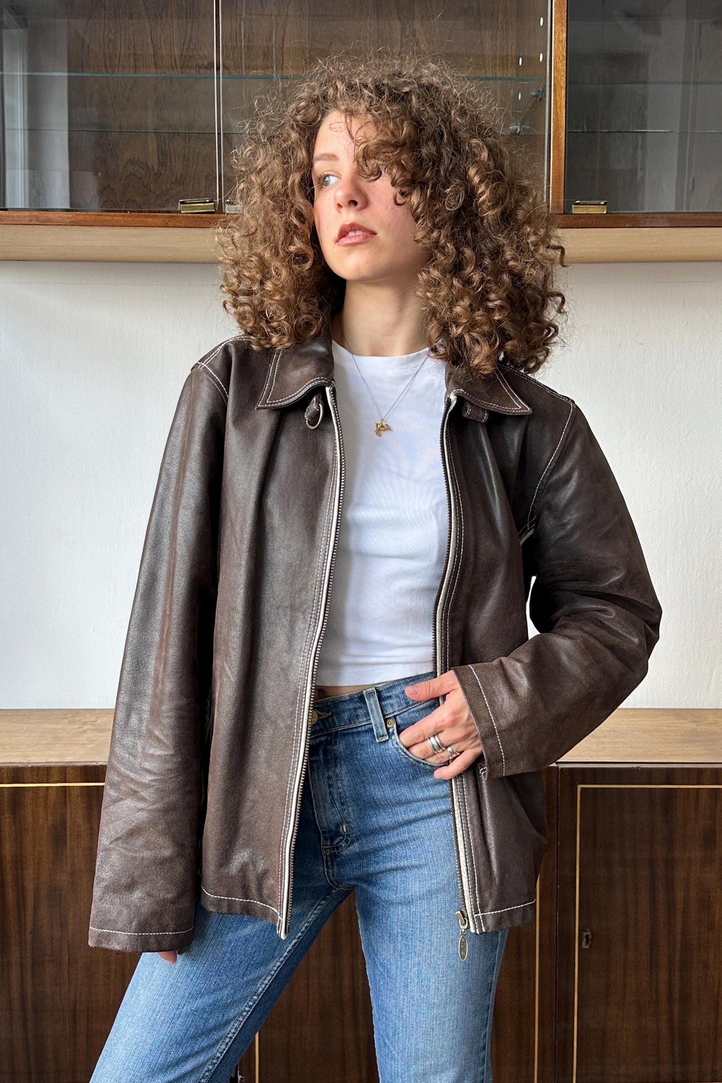 A young woman with curly hair wearing a brown leather jacket and blue jeans stands in front of a wooden cabinet.