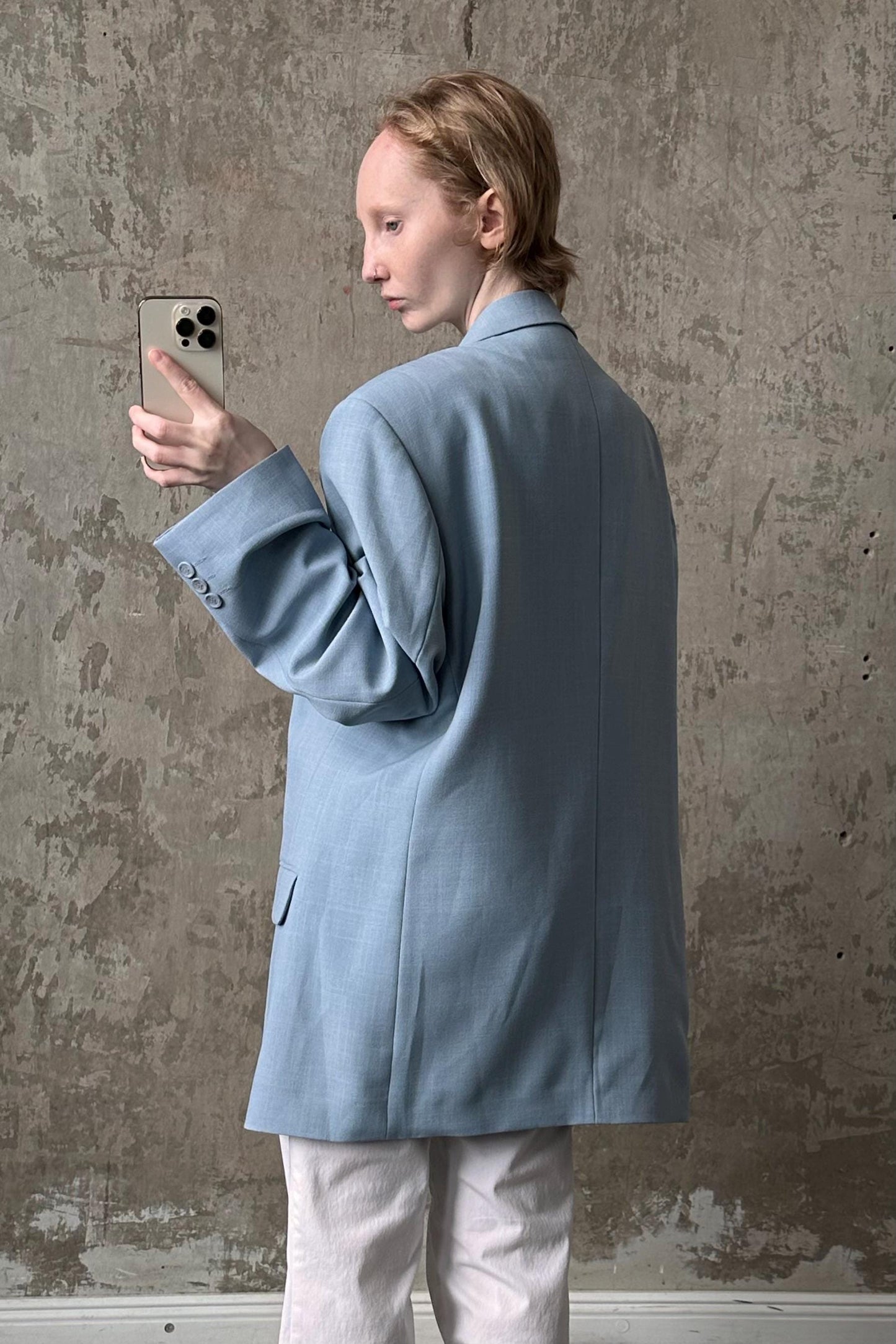 A person in a light blue blazer is holding a smartphone and looking at it, standing against a textured concrete wall.