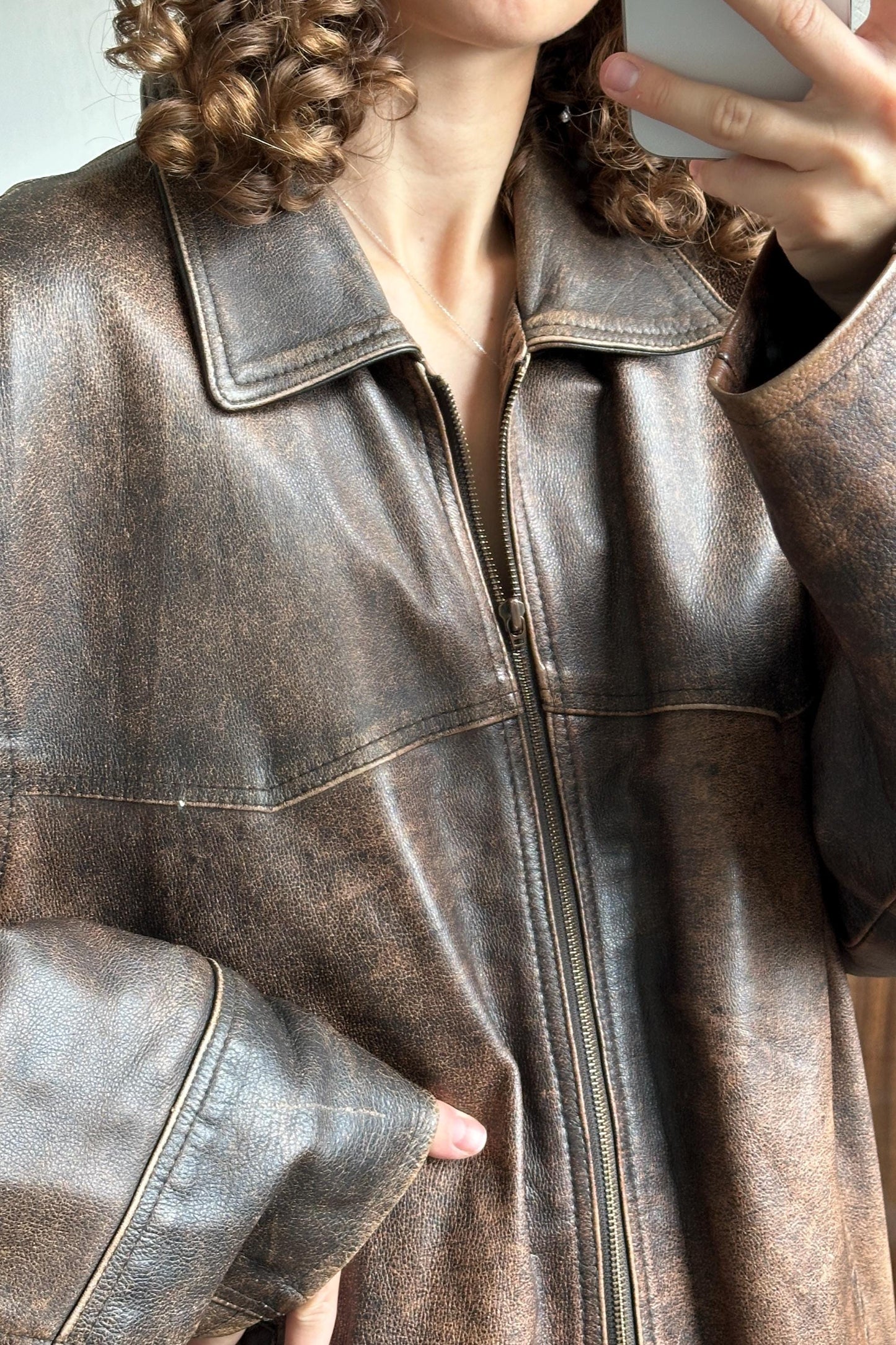 A close-up of a person wearing a brown leather jacket, taking a selfie with a phone.