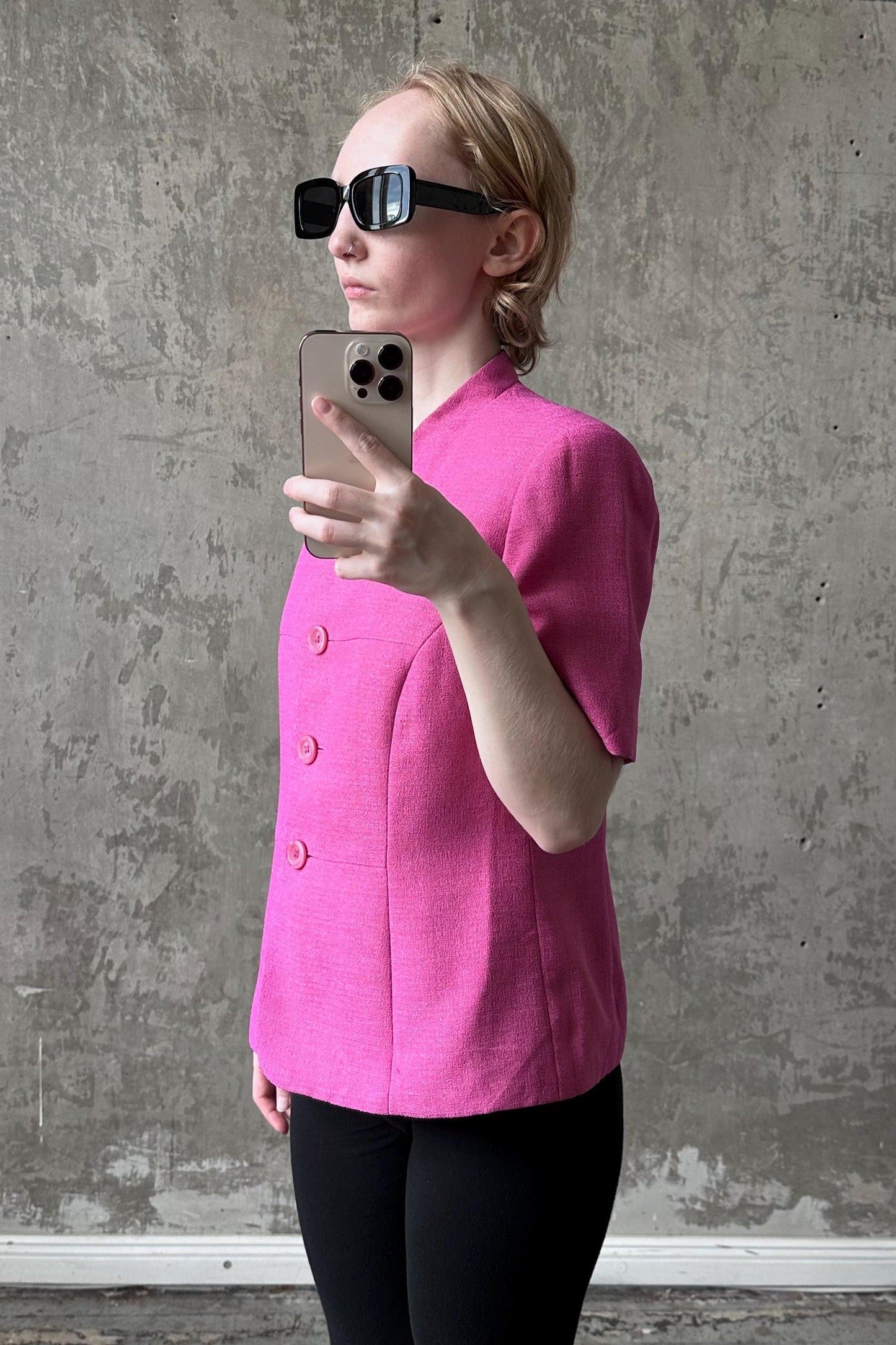 A woman wearing a pink blazer and sunglasses is taking a selfie with her iPhone against a concrete wall.