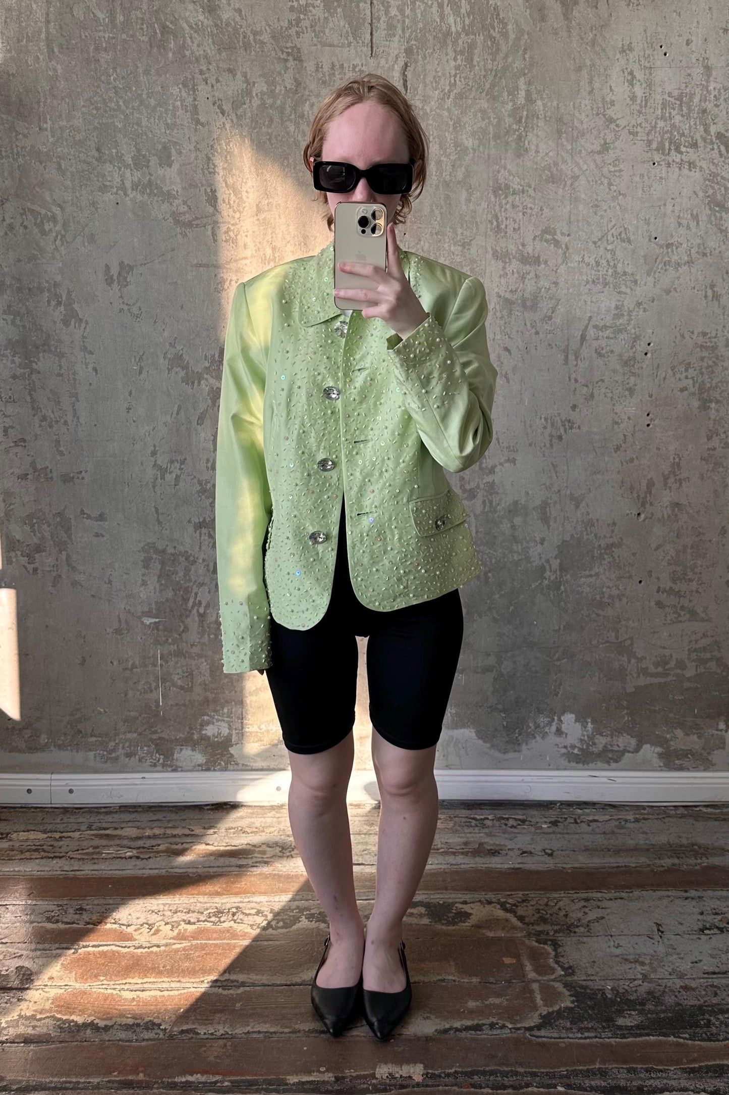 A person wearing a green jacket and black shorts is taking a selfie with a smartphone in front of a concrete wall.