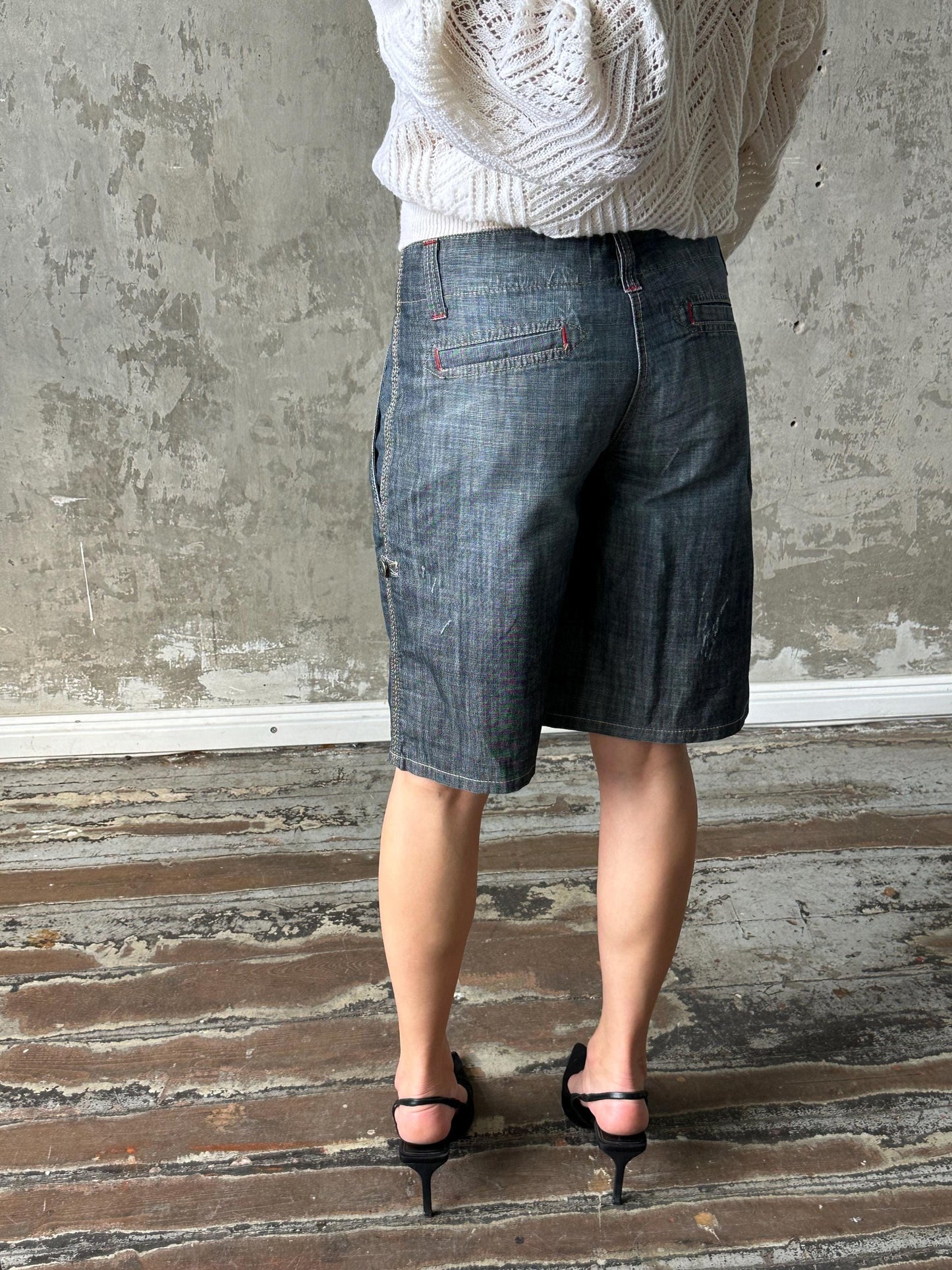 A person wearing a white sweater and black high heels stands in front of a concrete wall.