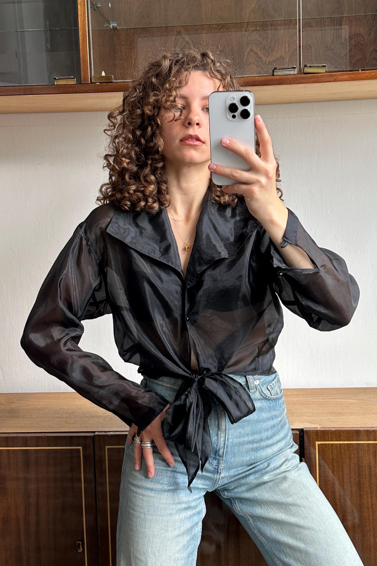 A woman with curly hair is taking a selfie with a smartphone, wearing a black blouse and blue jeans.