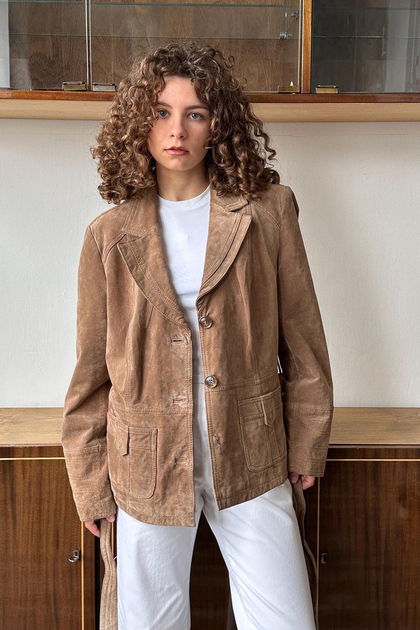 A young woman with curly hair wearing a tan jacket and white pants stands in front of a wooden cabinet.
