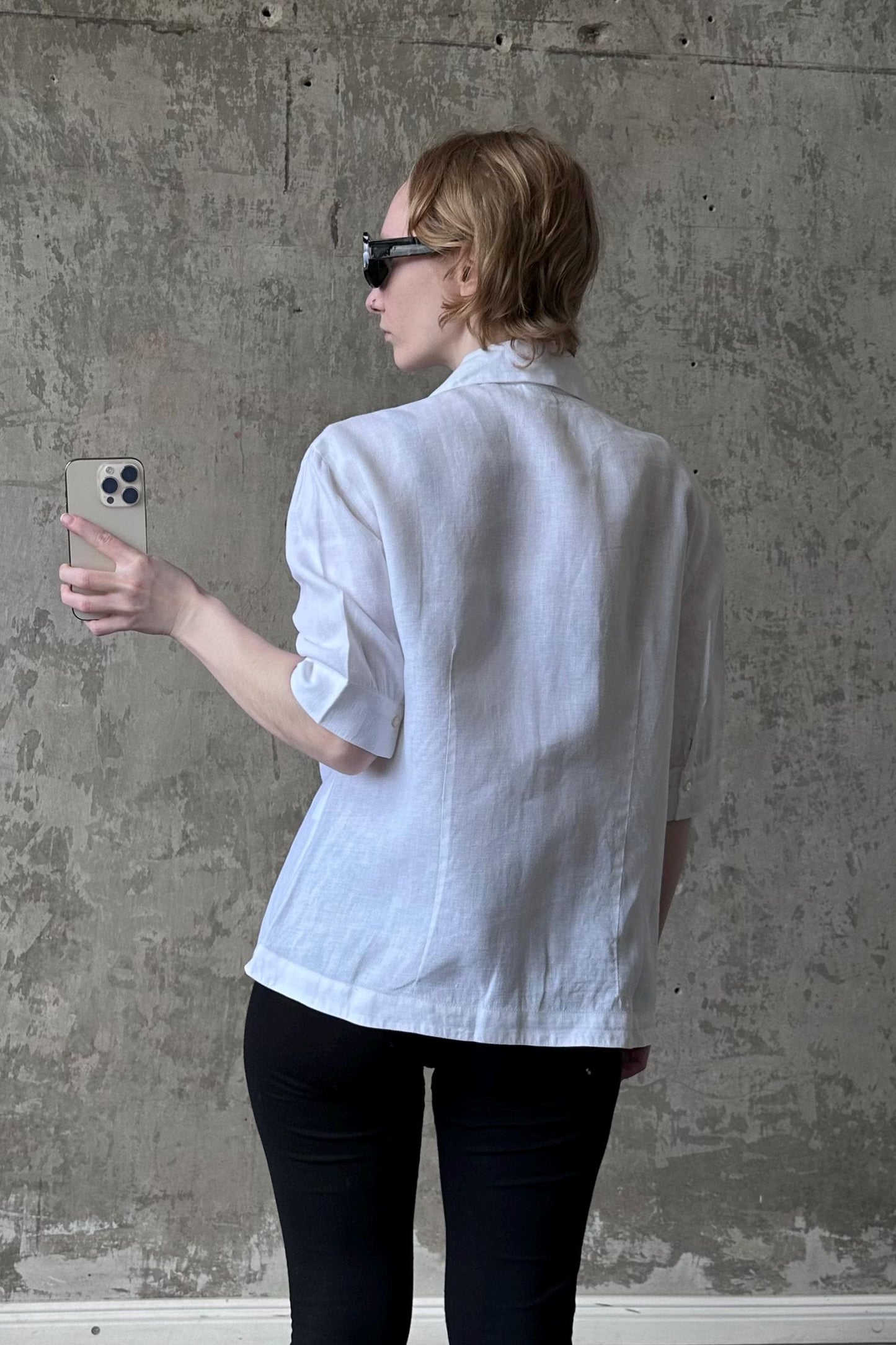 A person wearing a white shirt and sunglasses is holding a phone in front of a concrete wall.