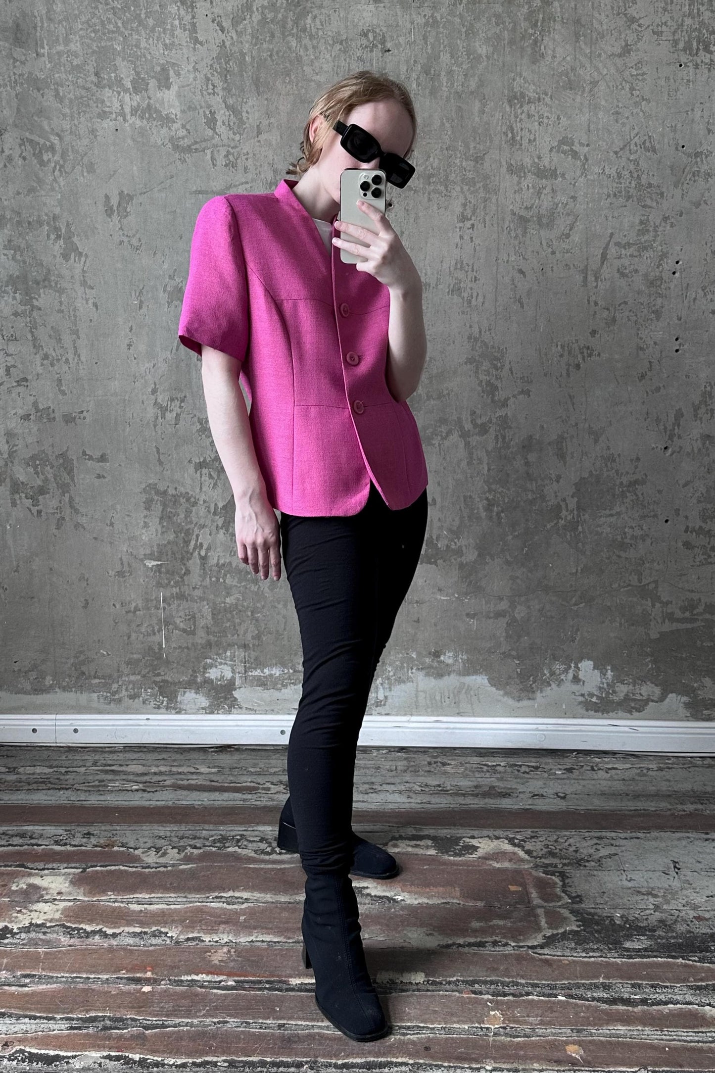 A person wearing a pink blazer and black pants is taking a selfie with a smartphone in front of a concrete wall.