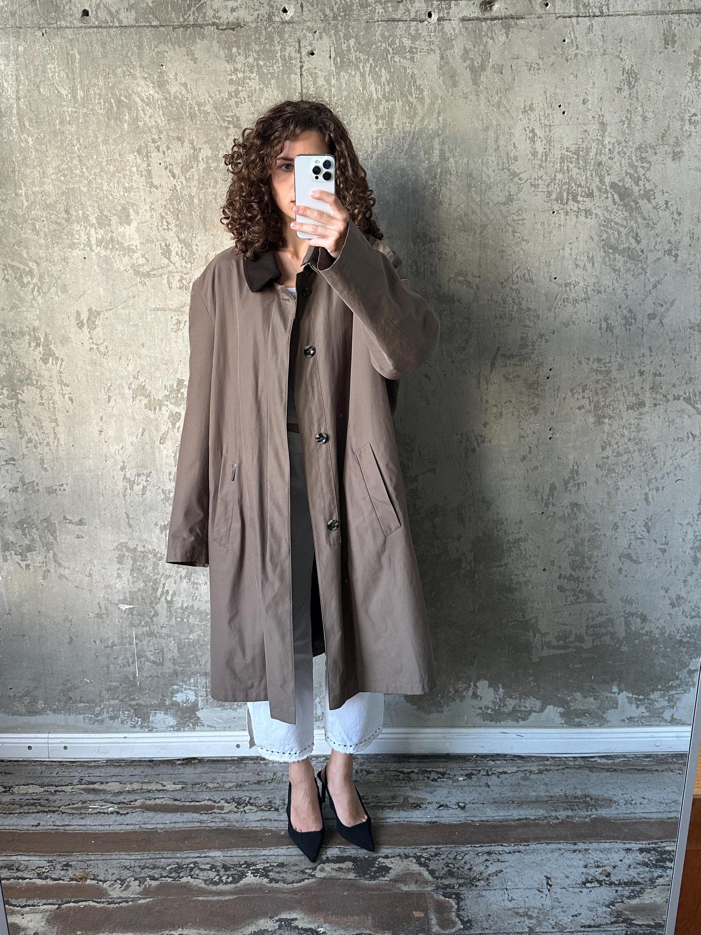 A woman in a brown coat is taking a selfie with her phone against a concrete wall.