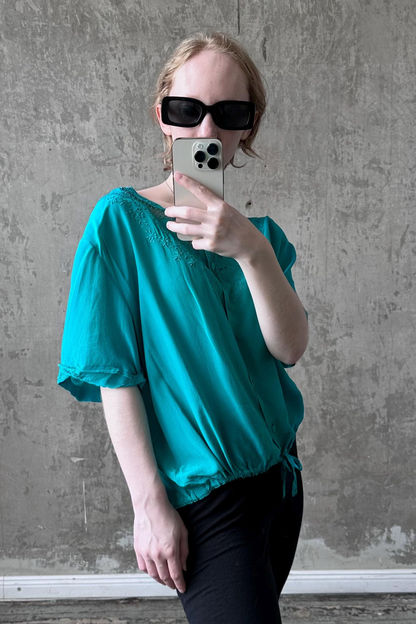 A woman wearing a teal blouse and sunglasses is taking a selfie with her iPhone against a concrete wall.