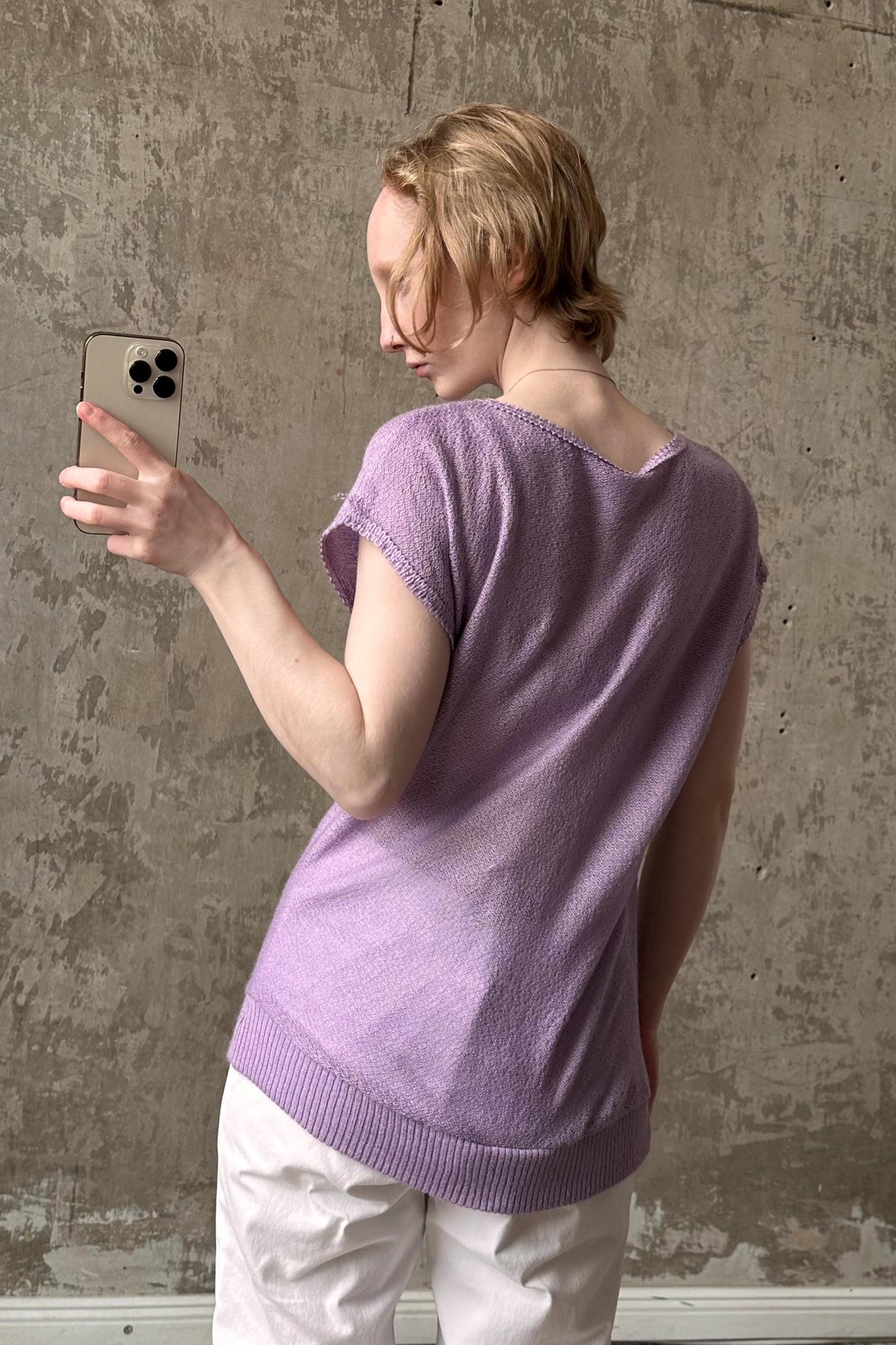 A woman in a purple sweater is taking a selfie with her iPhone against a concrete wall.