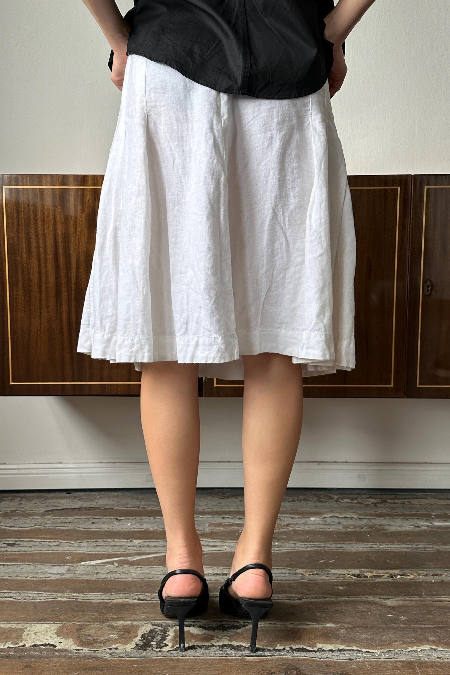 A person wearing a white skirt and black heels stands in front of a wooden cabinet.
