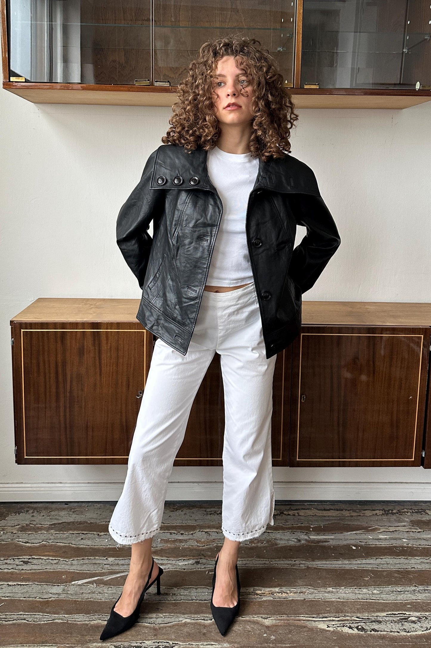 A woman with curly hair wearing a black leather jacket, white pants, and black heels stands in a room with wooden cabinets.