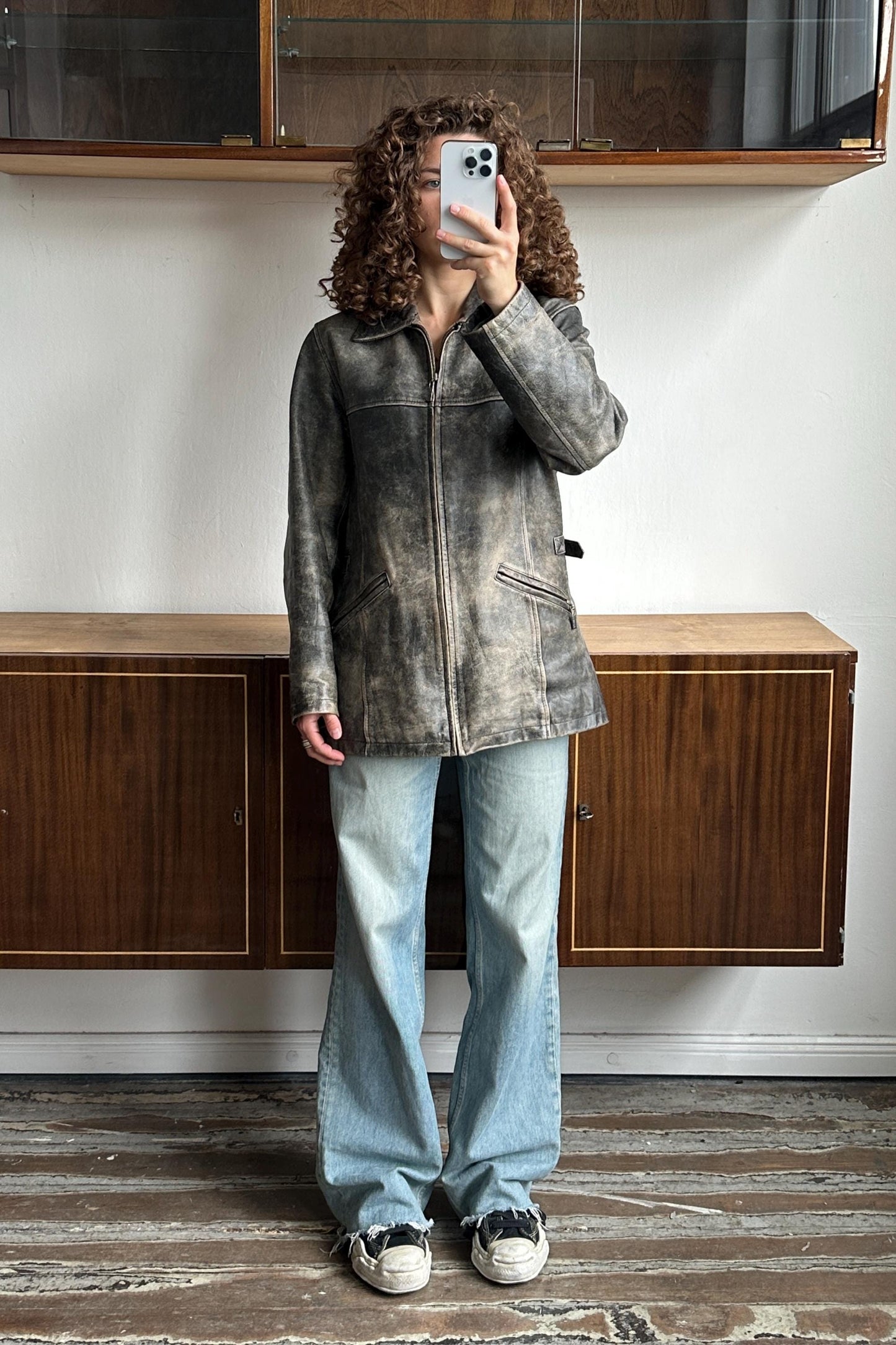 A person with curly hair is taking a selfie with a smartphone, standing in front of a wooden cabinet or shelf.