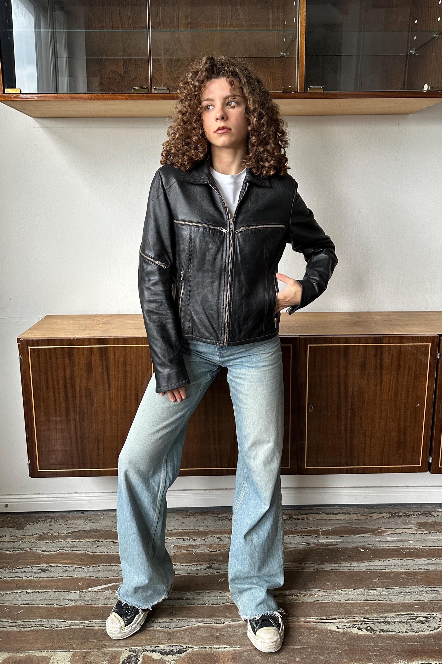 A young woman with curly hair wearing a black leather jacket and blue jeans stands in a room with wooden cabinets.