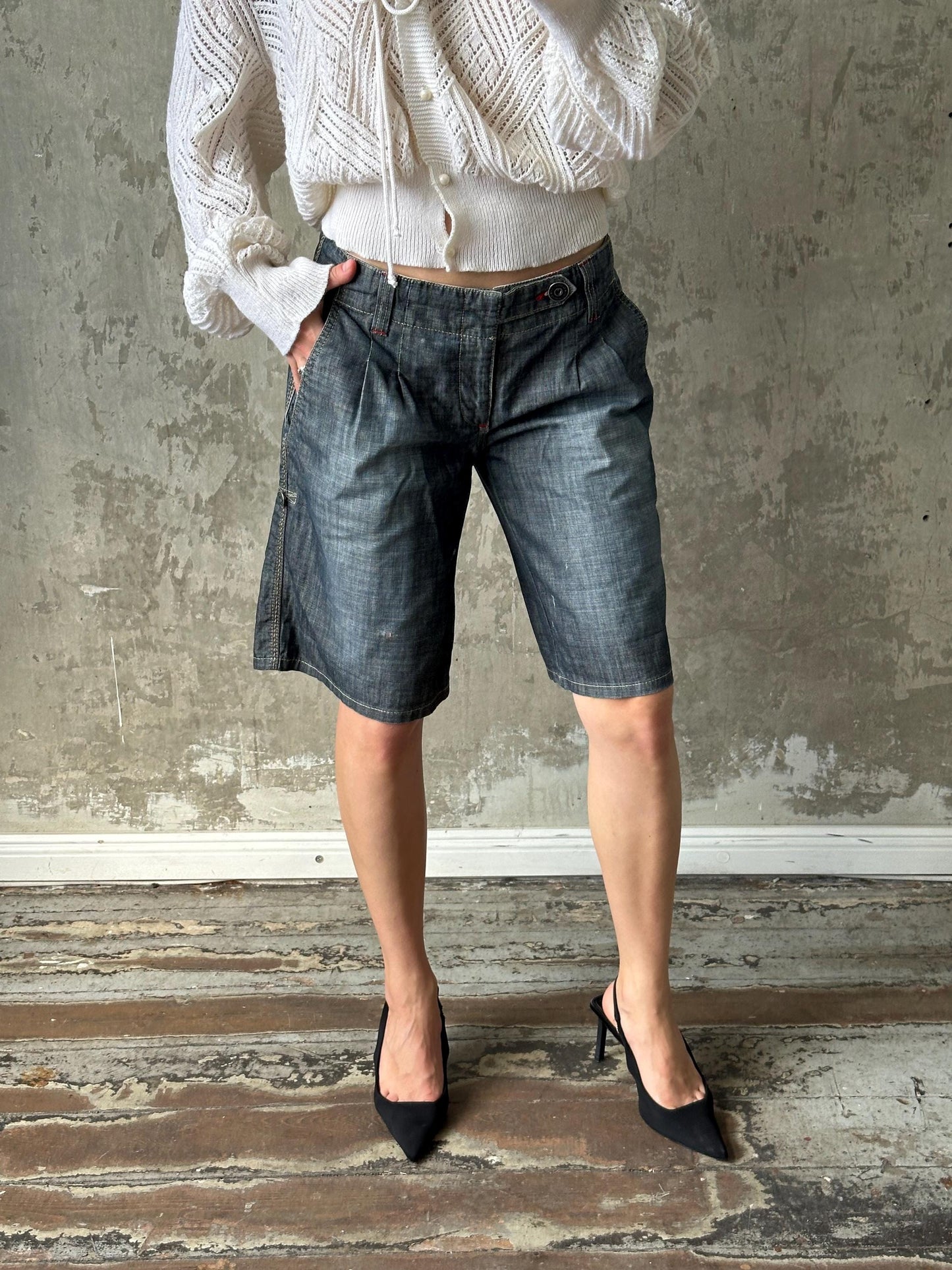 A person wearing a white sweater, blue jeans, and black high heels stands in front of a concrete wall.