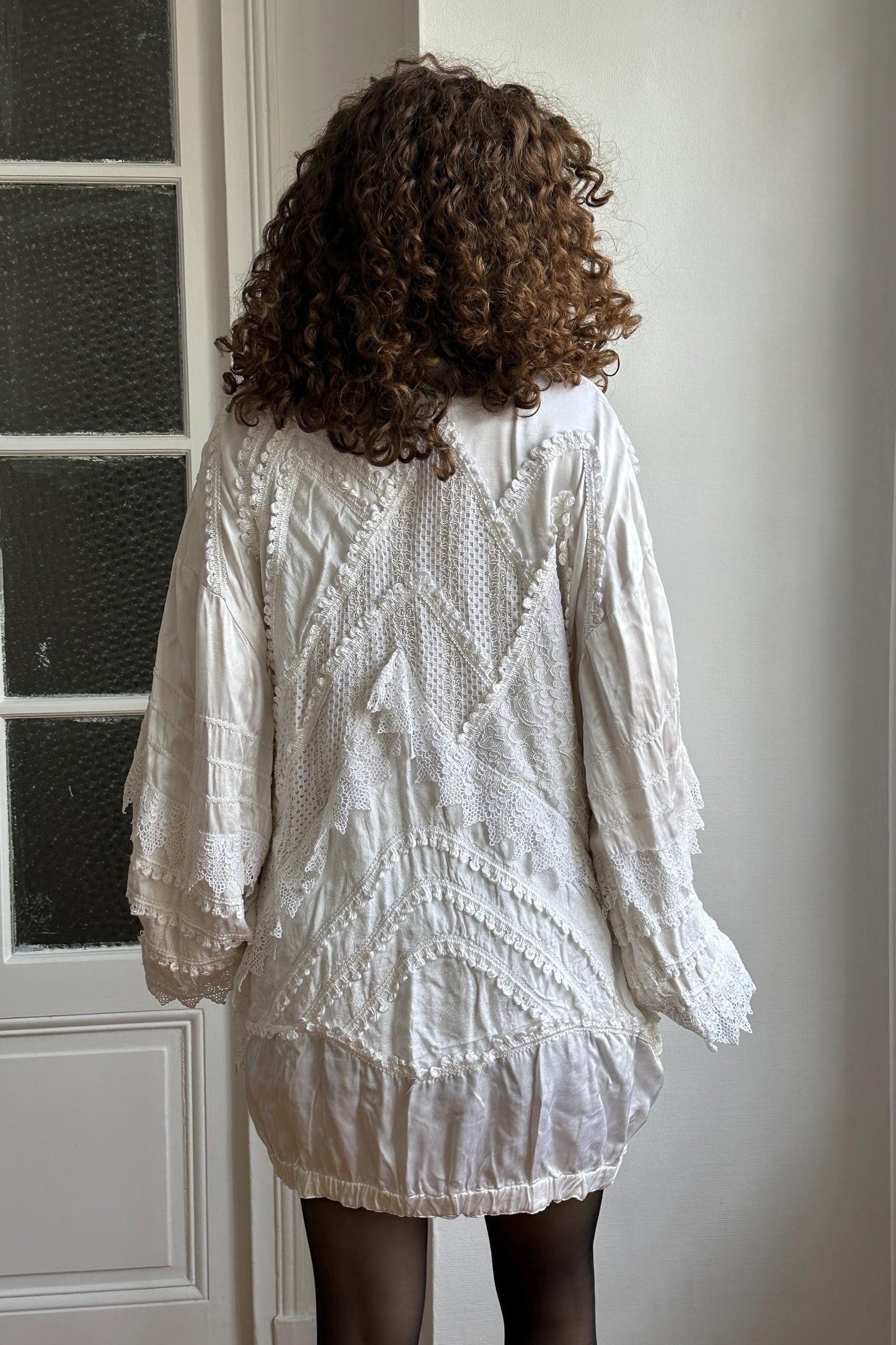 a person with curly hair wearing a white dress with intricate lace patterns and designs.