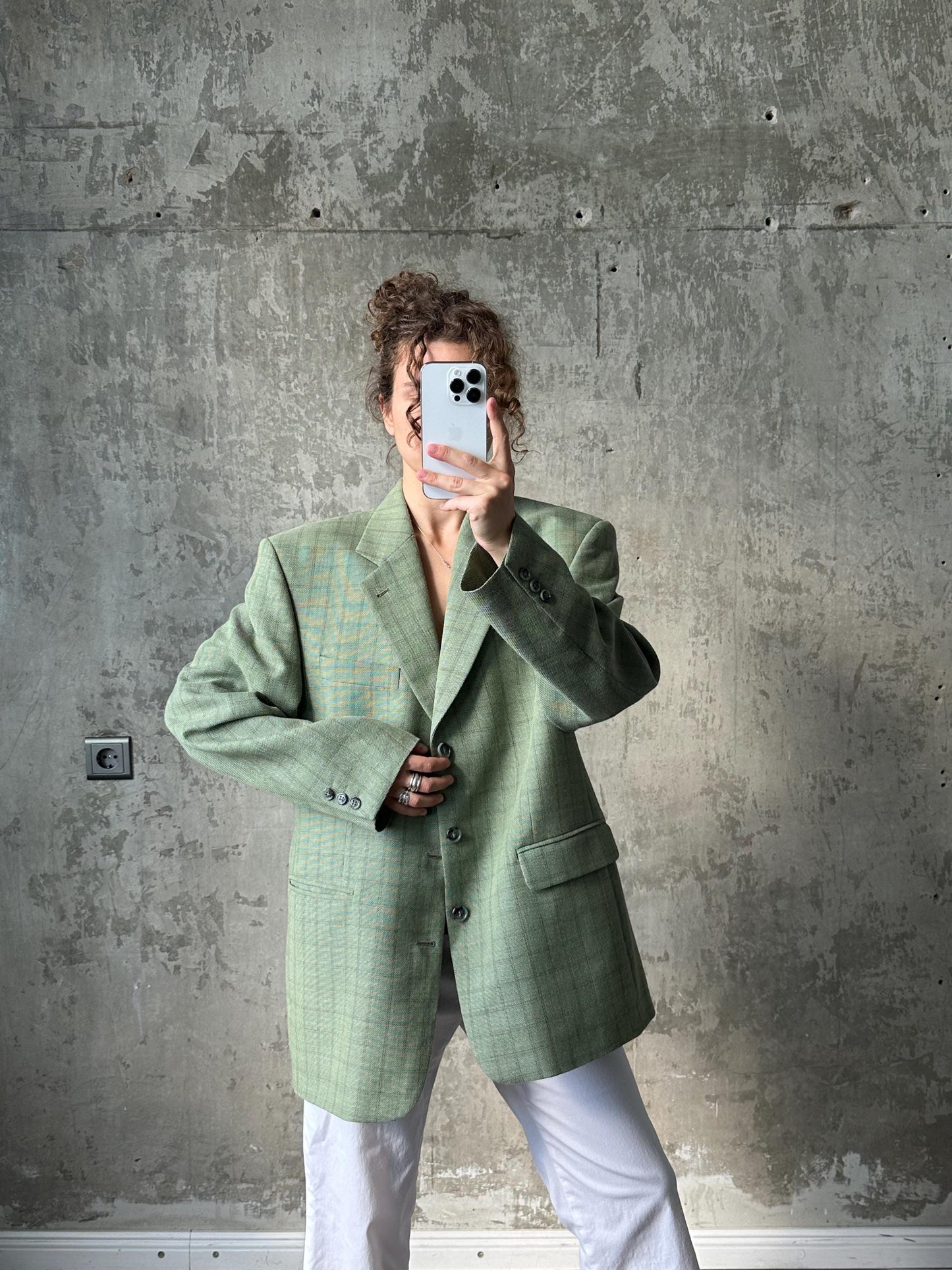 A person is taking a selfie with a white phone, wearing a green plaid jacket and white pants, against a concrete wall background.