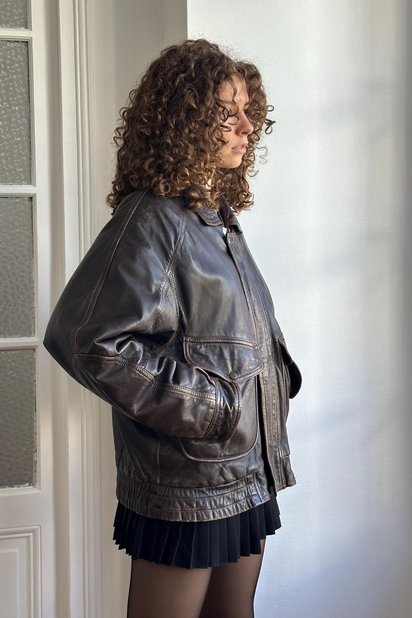 A woman with curly hair wearing a black leather jacket stands in front of a white wall, looking off to the side.