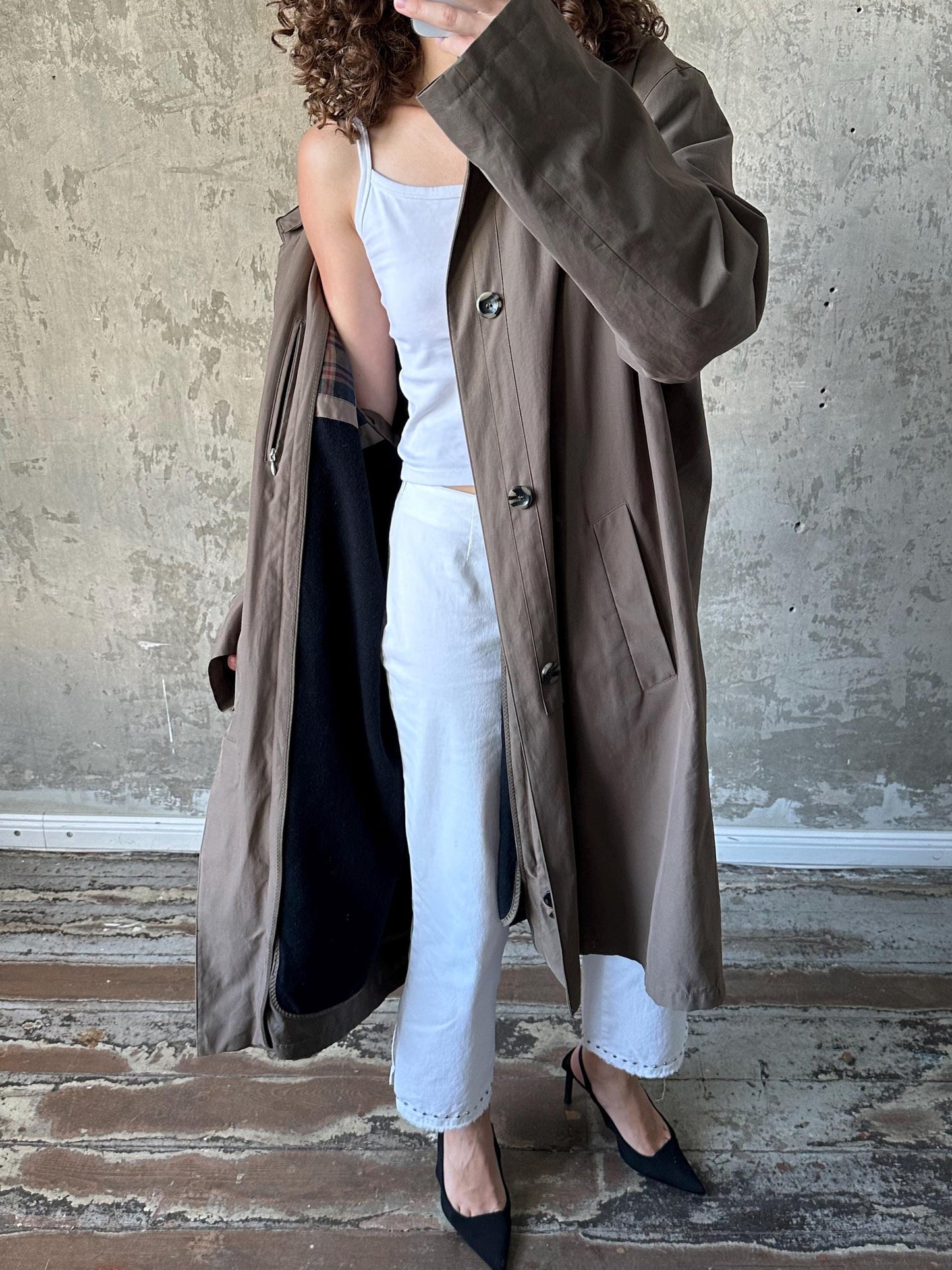 A person wearing a brown coat and white pants is standing in front of a concrete wall, with their arm raised and the coat partially unzipped.
