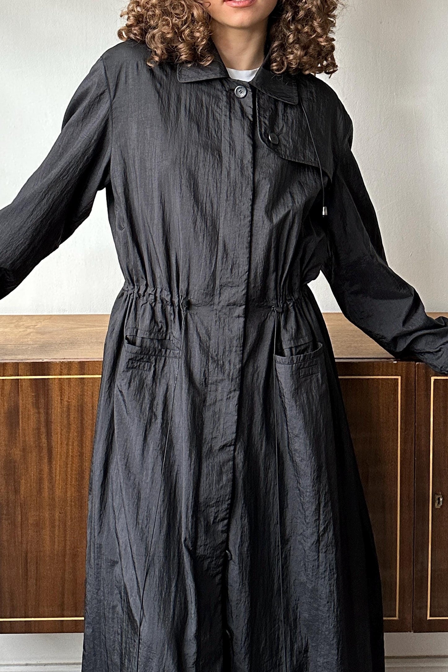 A person wearing a long, black coat with a button-down front and a drawstring hood.