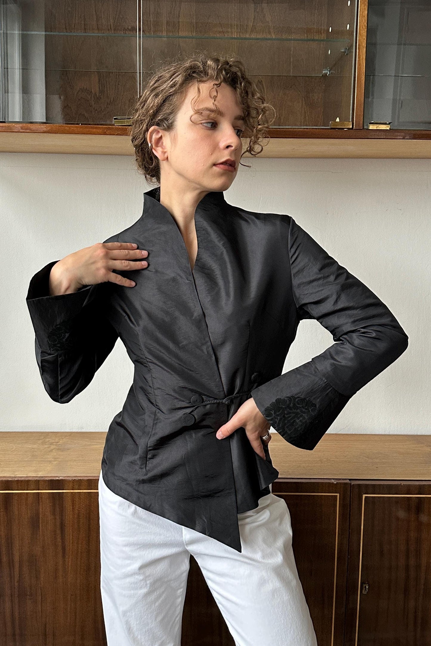 A woman wearing a black jacket stands in front of a wooden cabinet, with her hand on her hip.