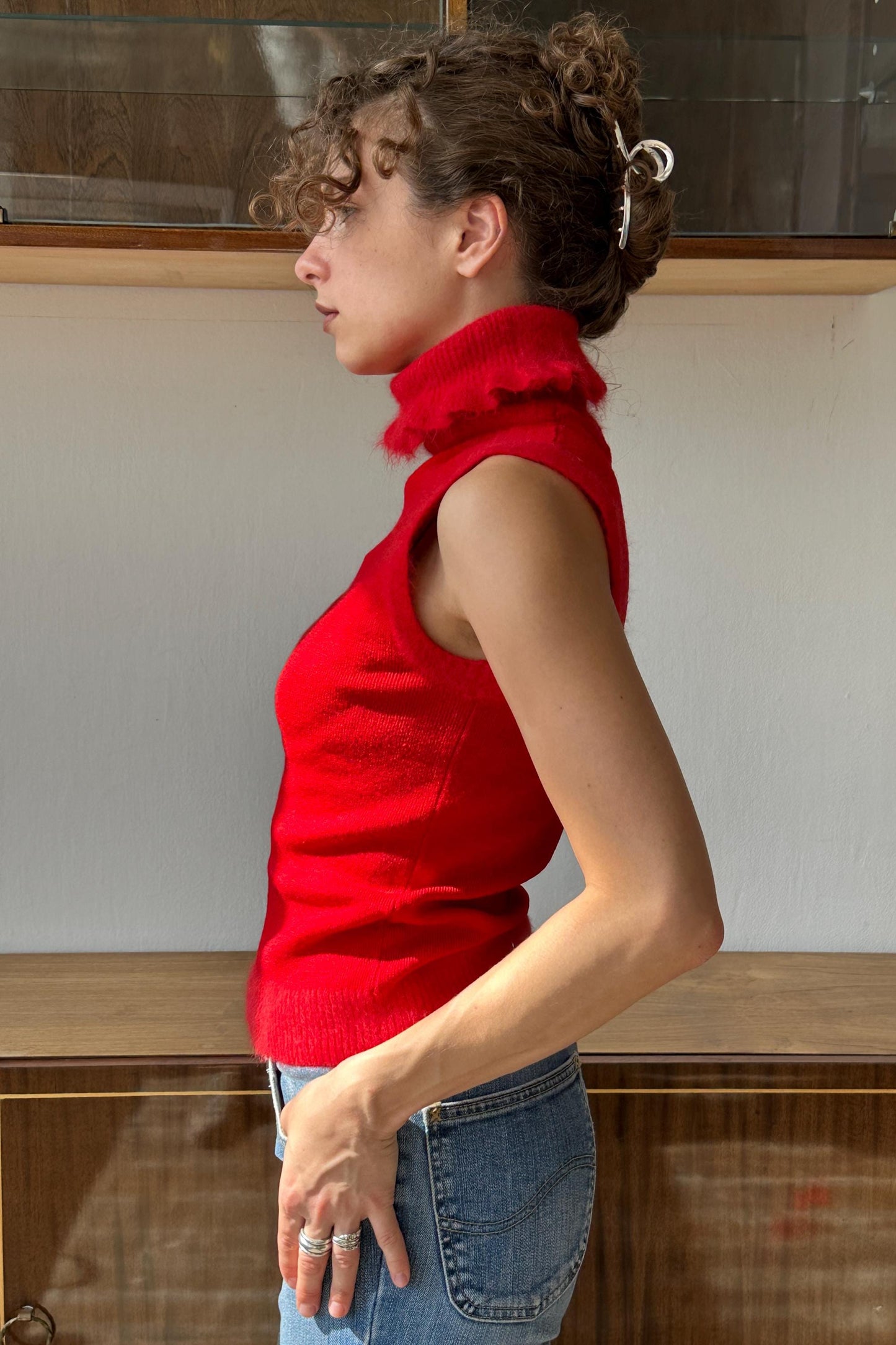 A woman wearing a red sleeveless top and jeans stands in front of a wooden cabinet.