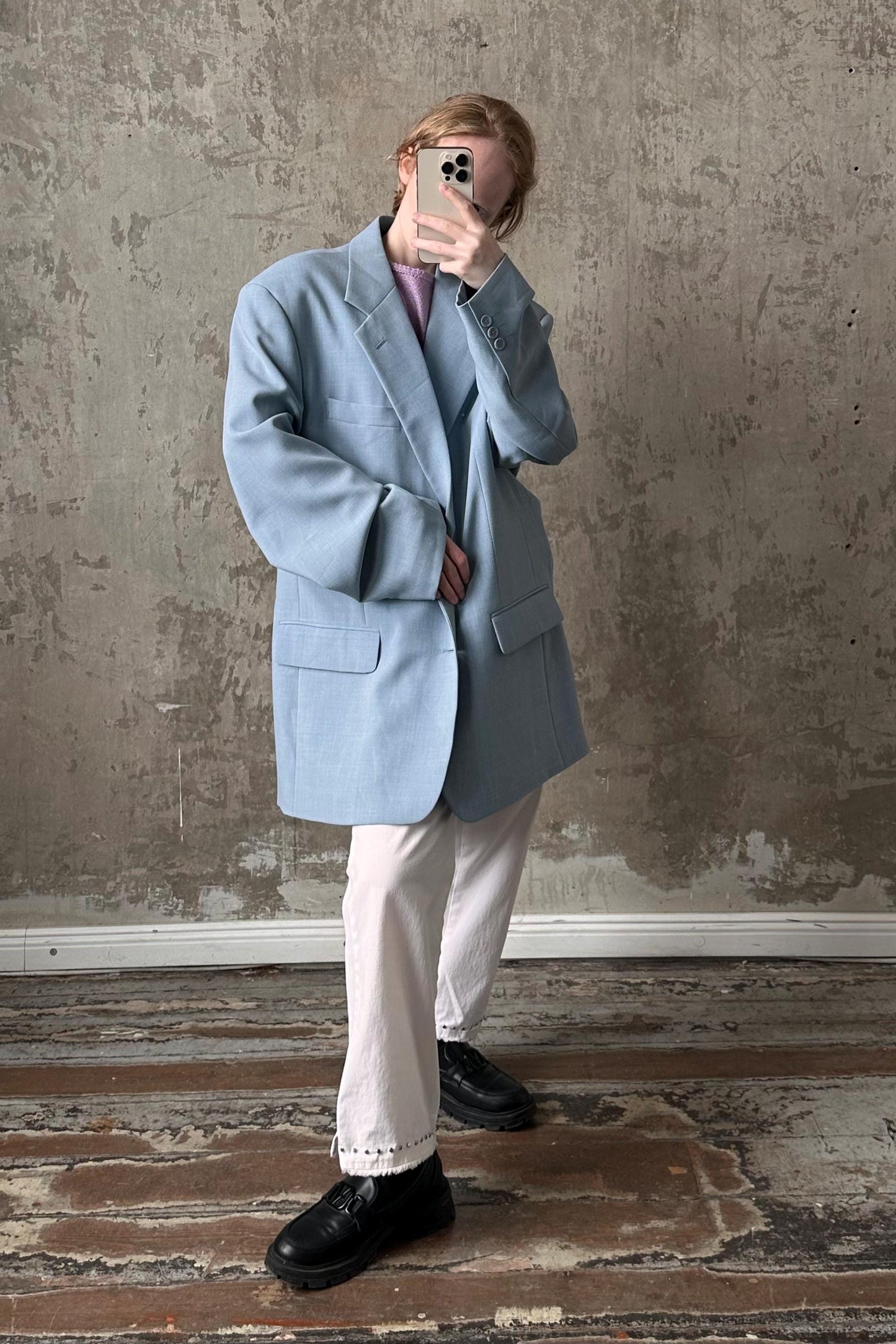 A person is taking a selfie with a smartphone, wearing a light blue blazer and white pants, standing in front of a concrete wall.